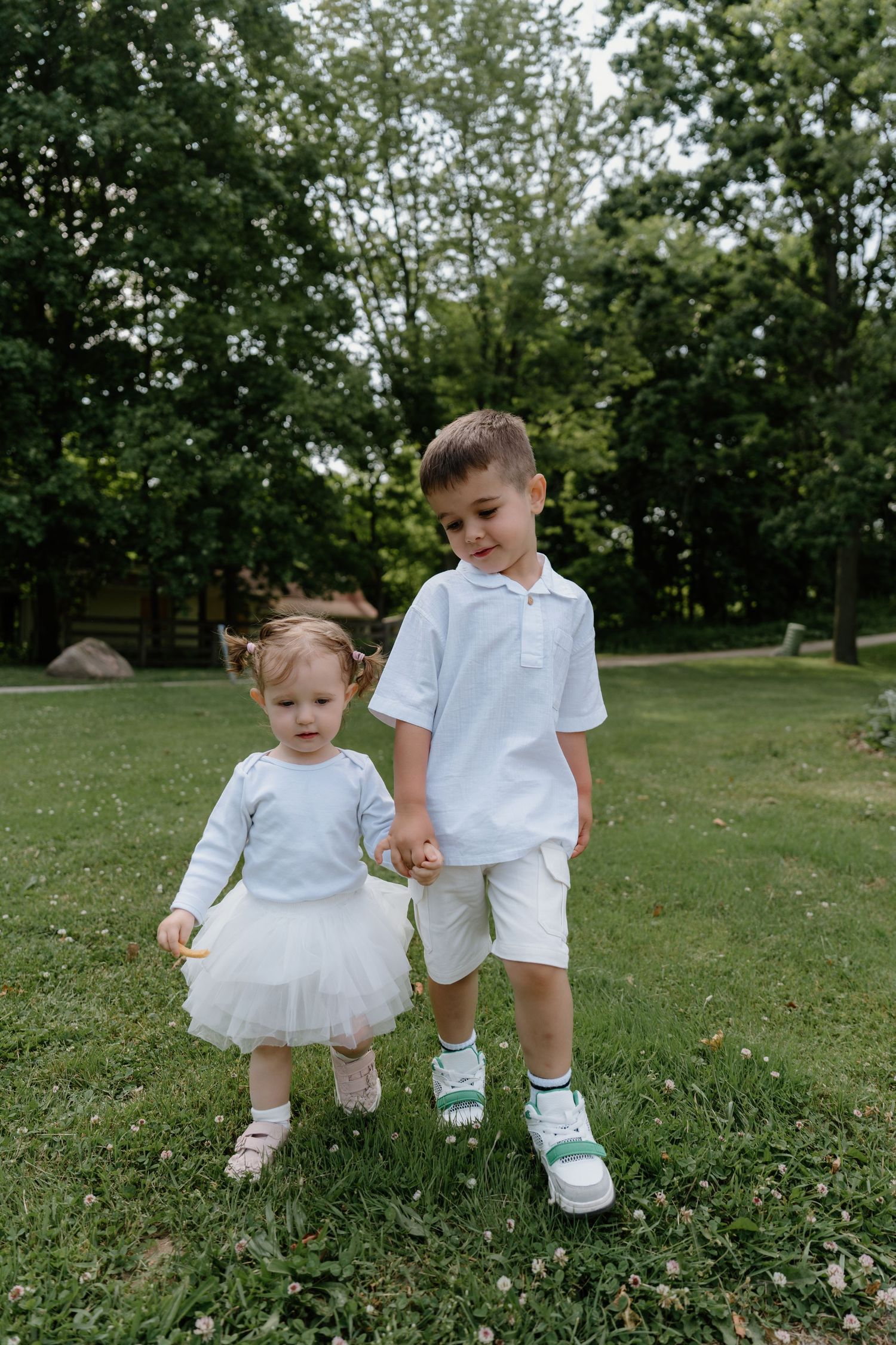 Two young children in white outfits hold hands while standing together on grass with trees in the background.