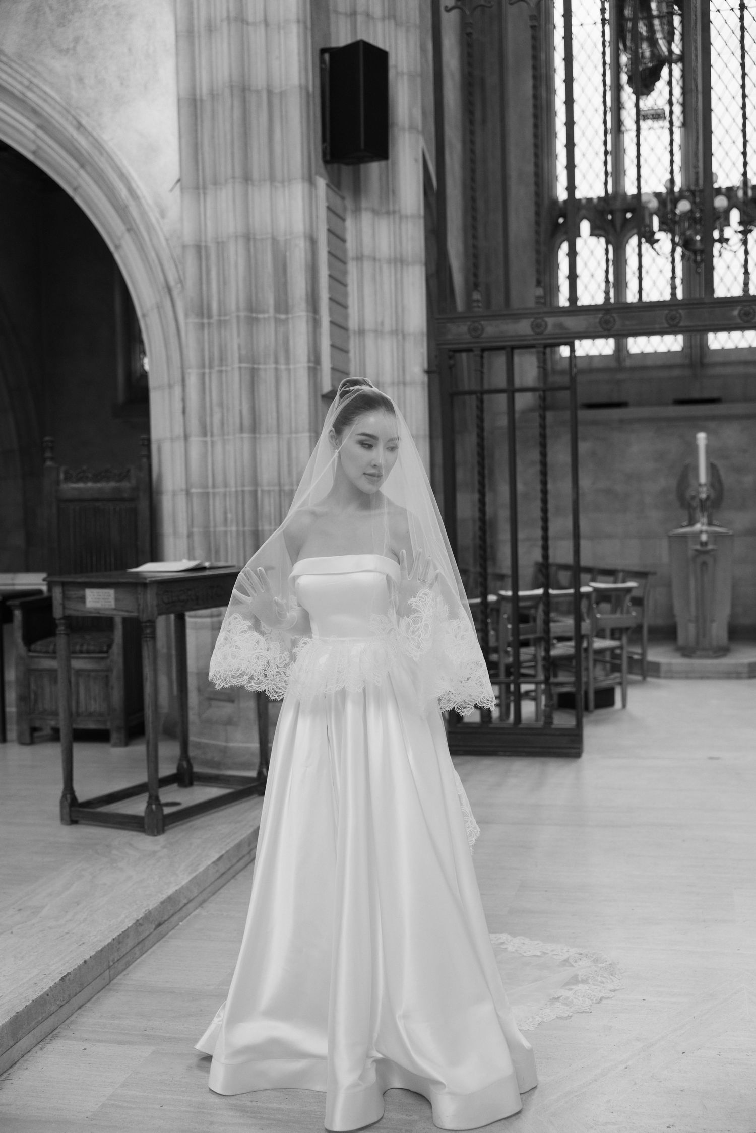 A bride in an elegant white satin wedding gown with lace veil stands in a cathedral with gothic architectural details.