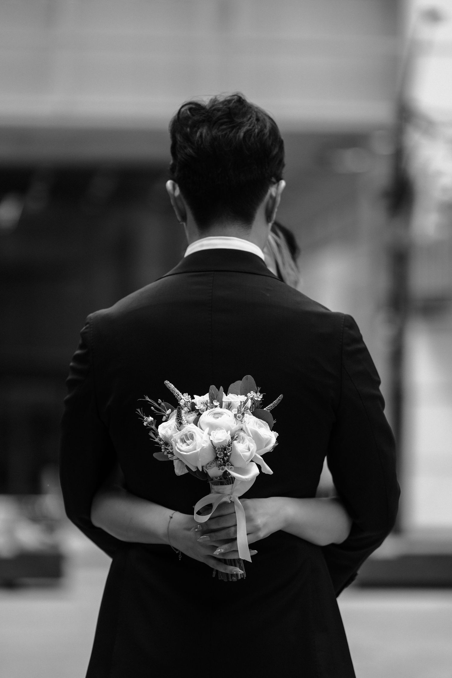 Back view of groom in black suit holding delicate white bouquet behind his back in black and white photography.