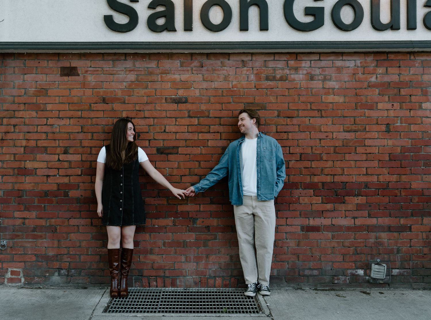 Two people stand against a red brick wall beneath a storefront sign while holding hands and looking at each other.