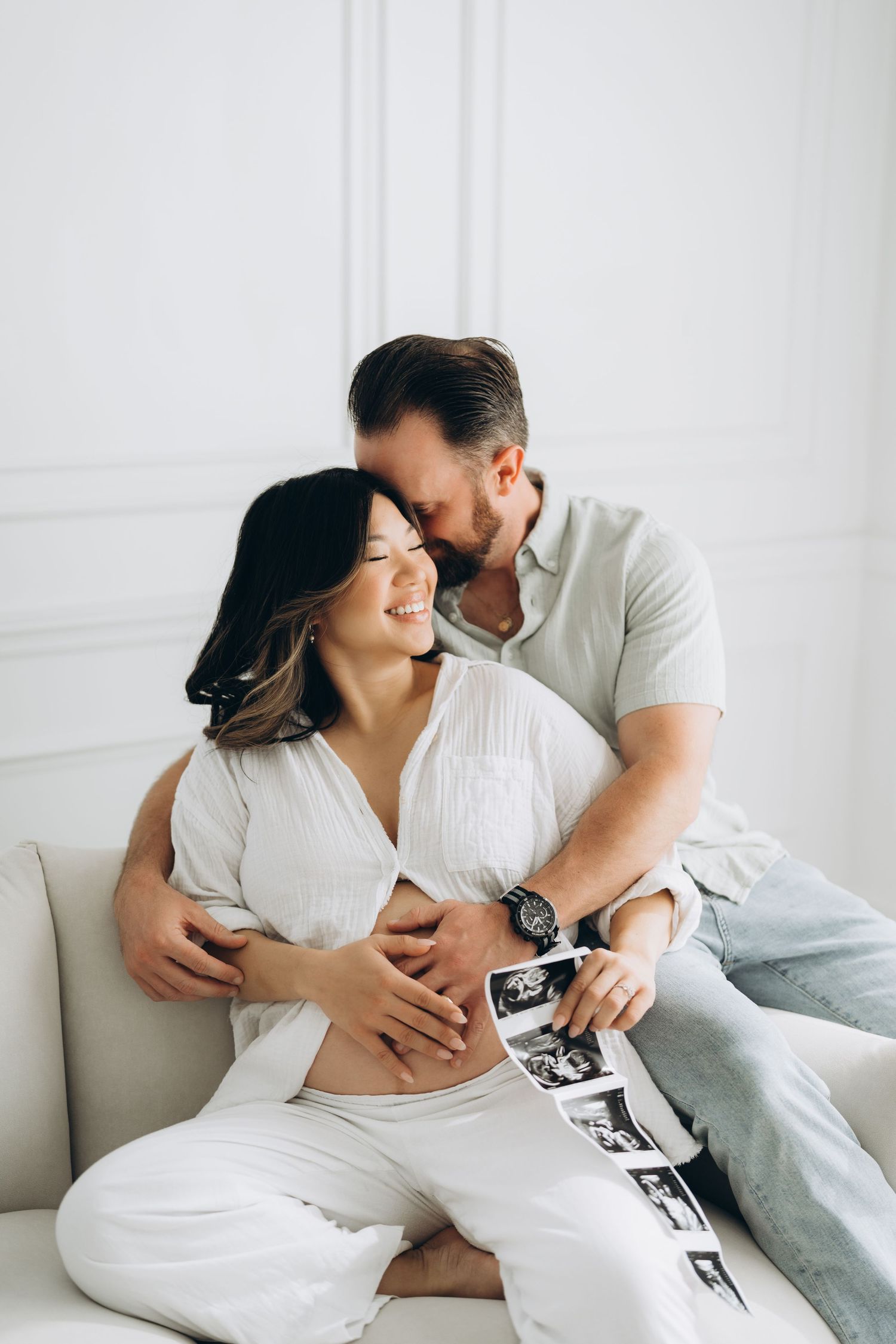 A couple dressed in white lounges intimately together on a light colored couch in a bright minimalist setting.
