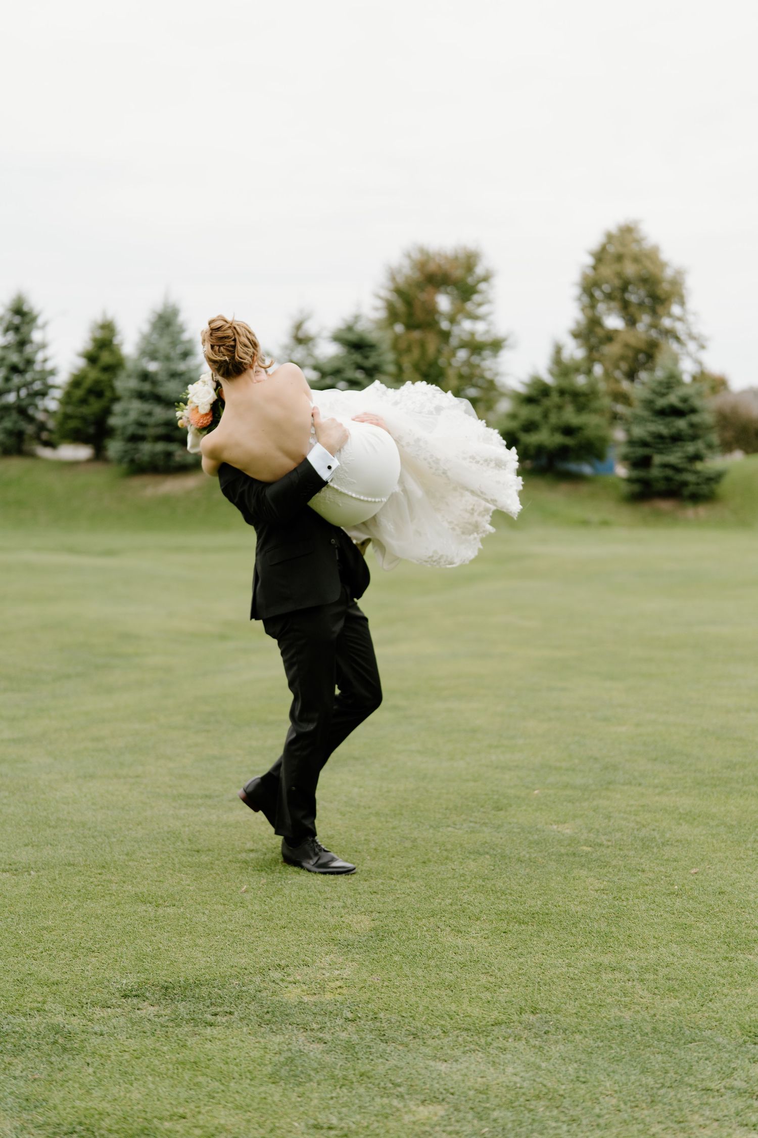 A joyful wedding couple embraces on a green golf course with evergreen trees in the background.