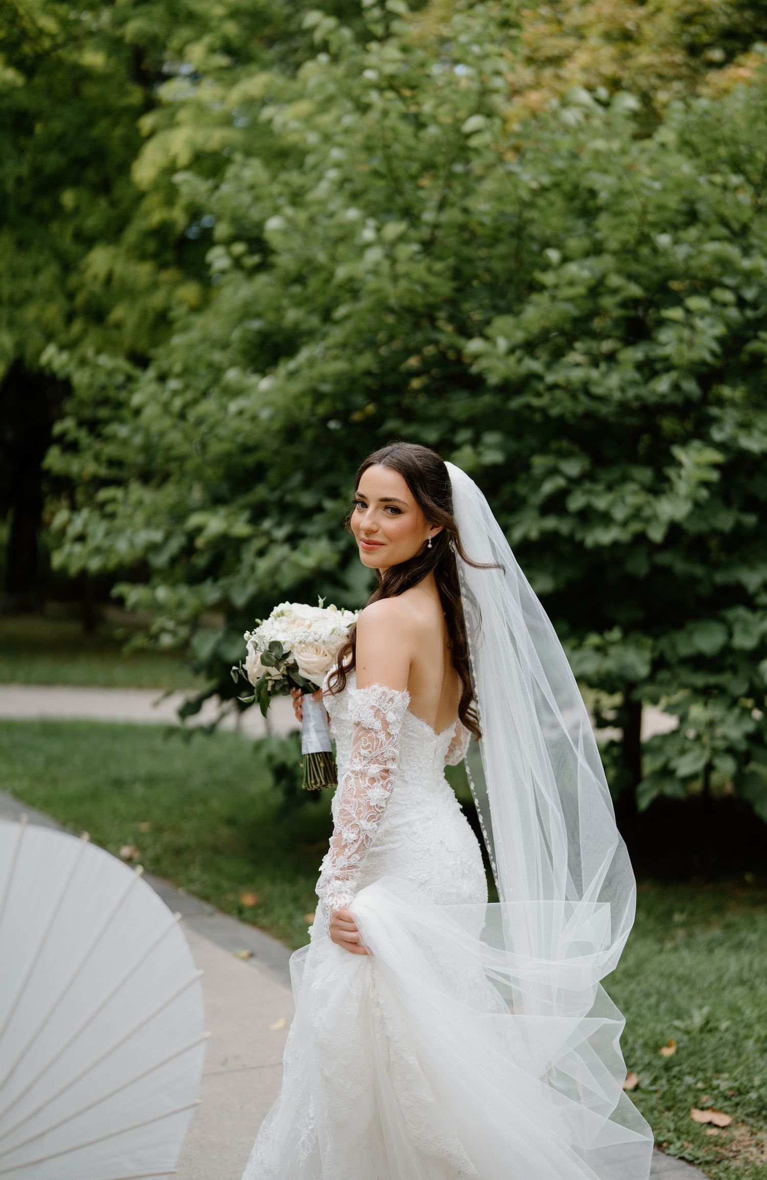 A bride in a flowing white gown with floral details poses gracefully in a lush garden setting.