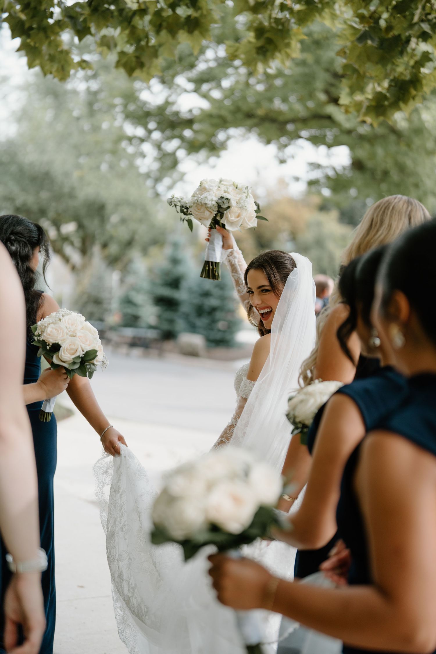 Bridesmaids and bride laugh joyfully together while holding white floral bouquets during outdoor wedding celebration.
