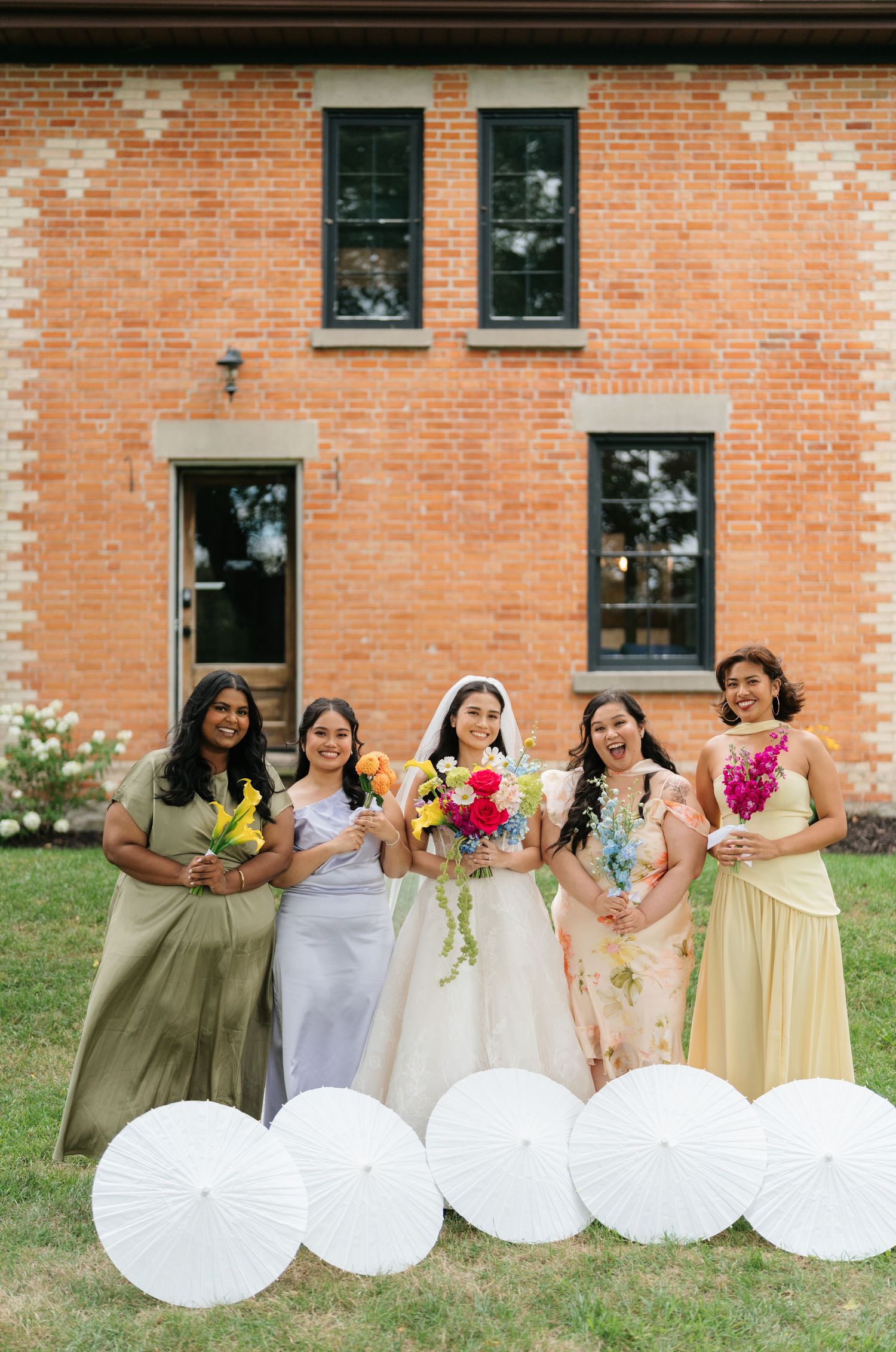Wedding party members hold white parasols while standing in a row against a brick building backdrop.