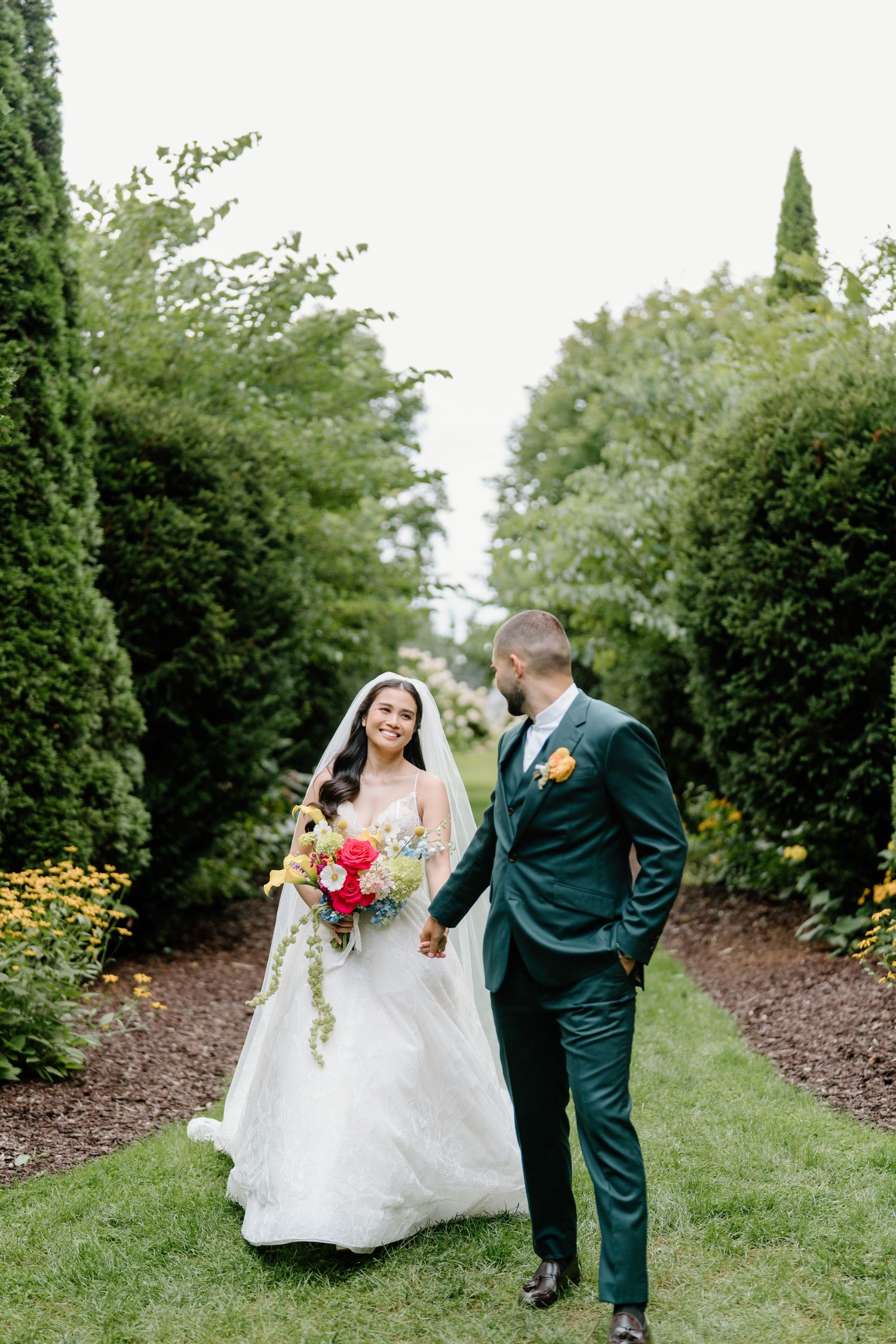 A bride in a white gown and groom in emerald green suit walk together on a garden path surrounded by tall hedges.