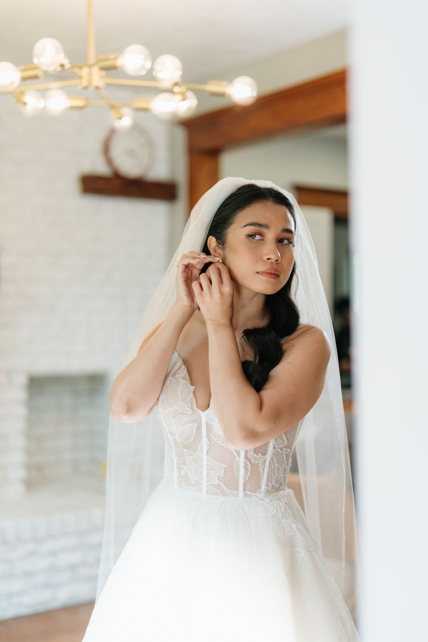 A bride adjusts her veil while standing in a light-filled room with modern decor and exposed wooden beams.