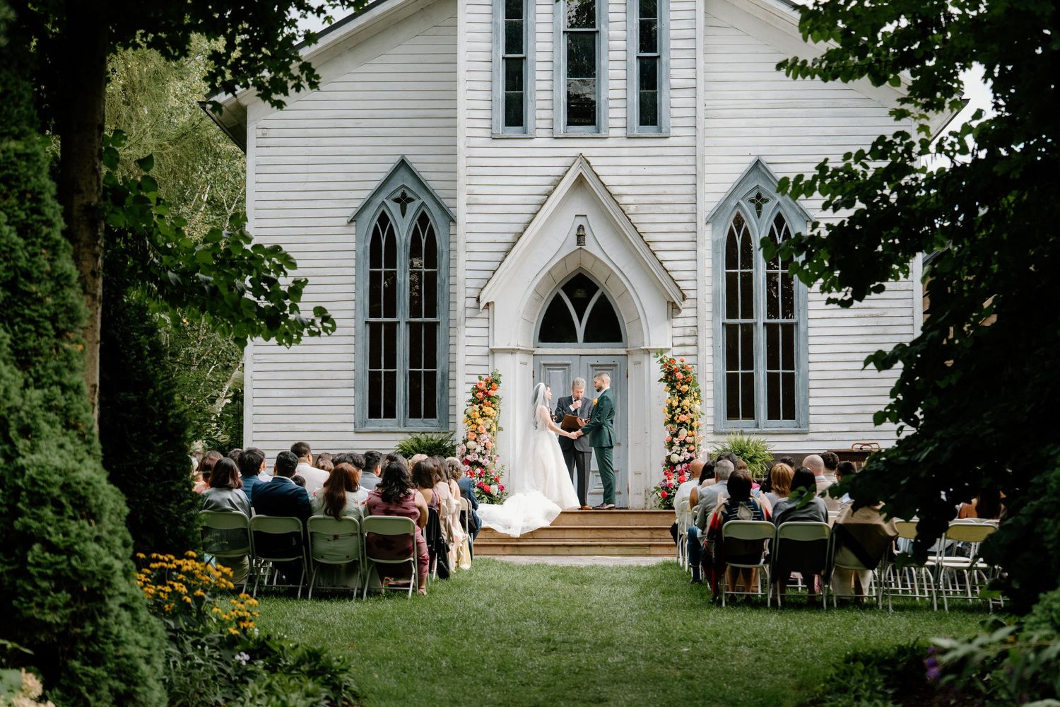 A beautiful outdoor wedding ceremony at a white country church with guests seated on wooden chairs in a garden setting.