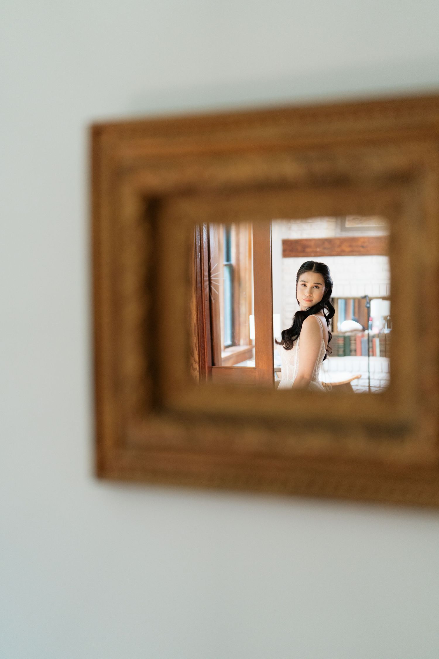 A reflection in a wooden framed mirror shows a figure in an interior space with soft natural lighting.
