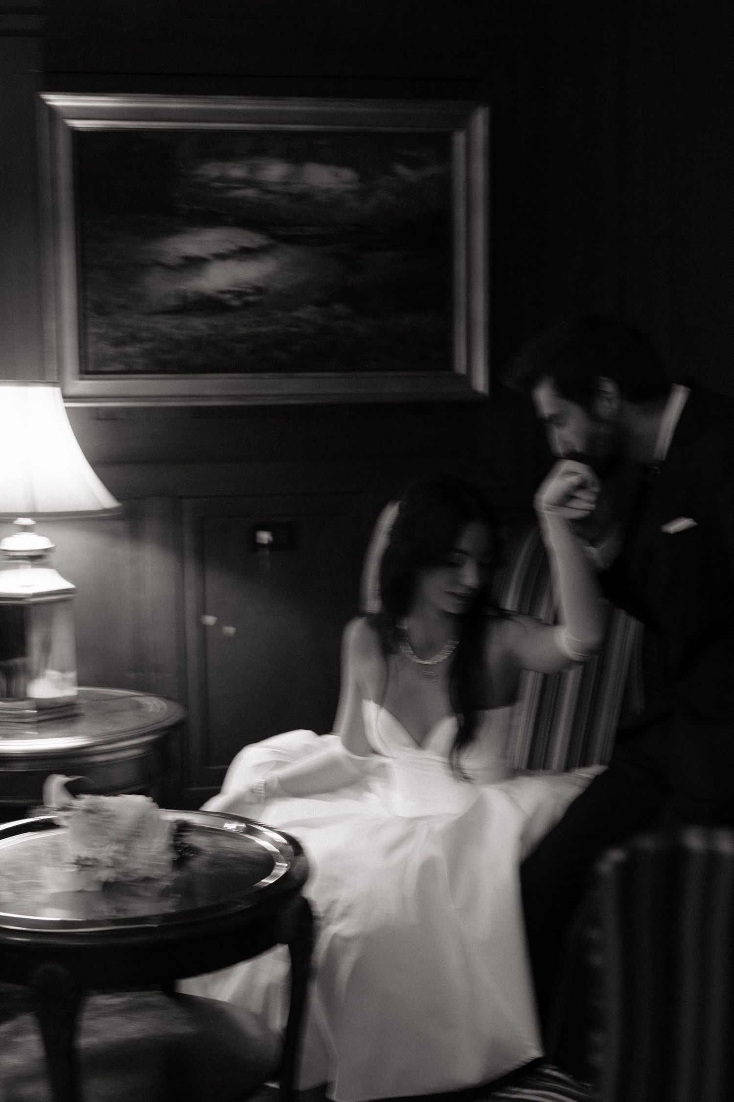 A black and white photograph showing a couple in formal attire sitting in a dimly lit room with vintage furniture.