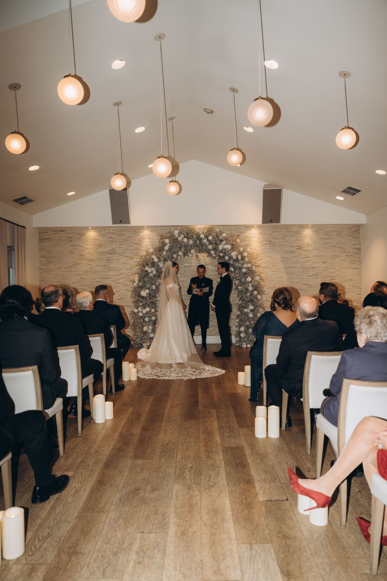 Indoor wedding ceremony with guests seated in white chairs facing a floral arch backdrop on light hardwood floors.
