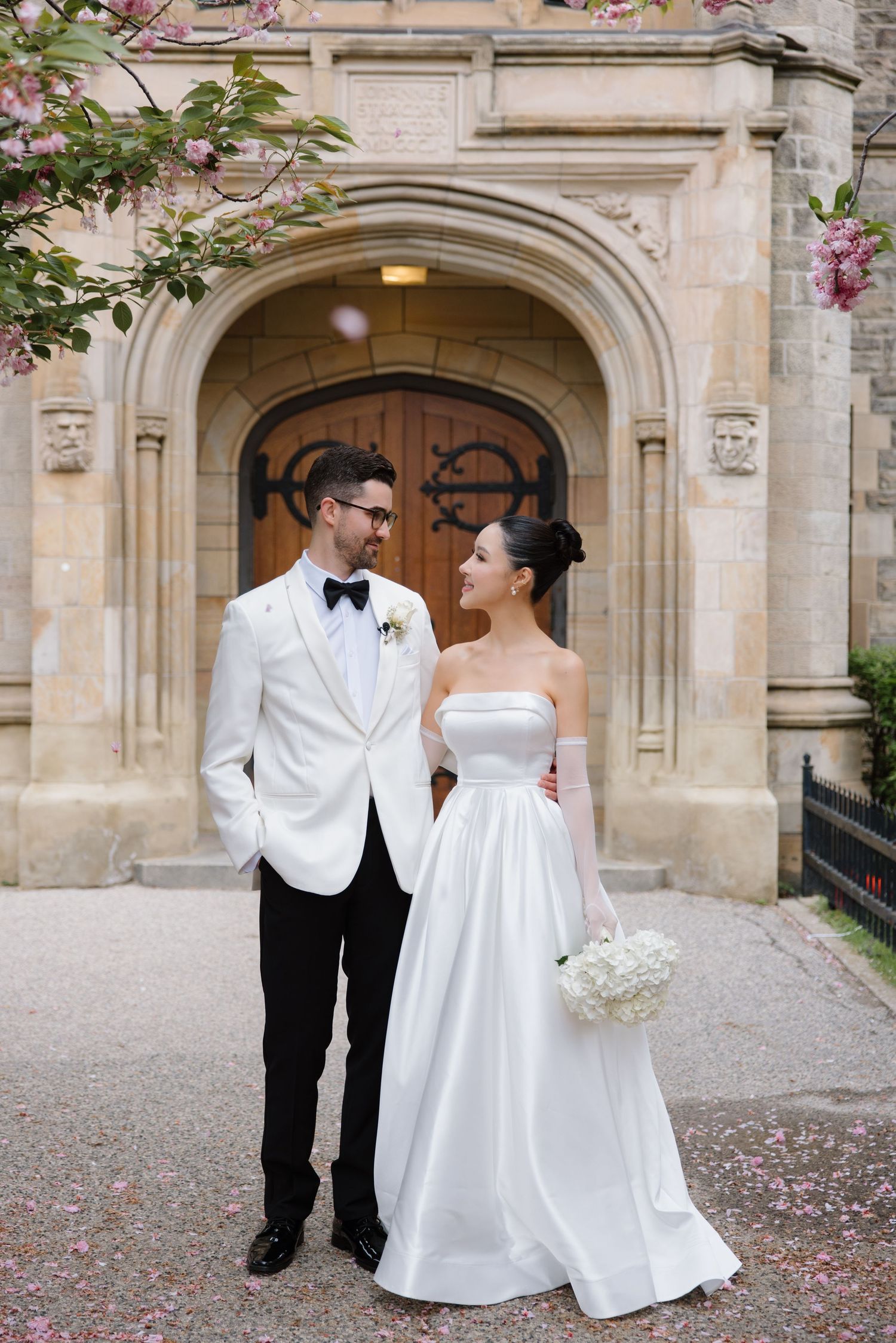 A couple in formal wedding attire stand together smiling in front of a stone archway with blooming flowers.