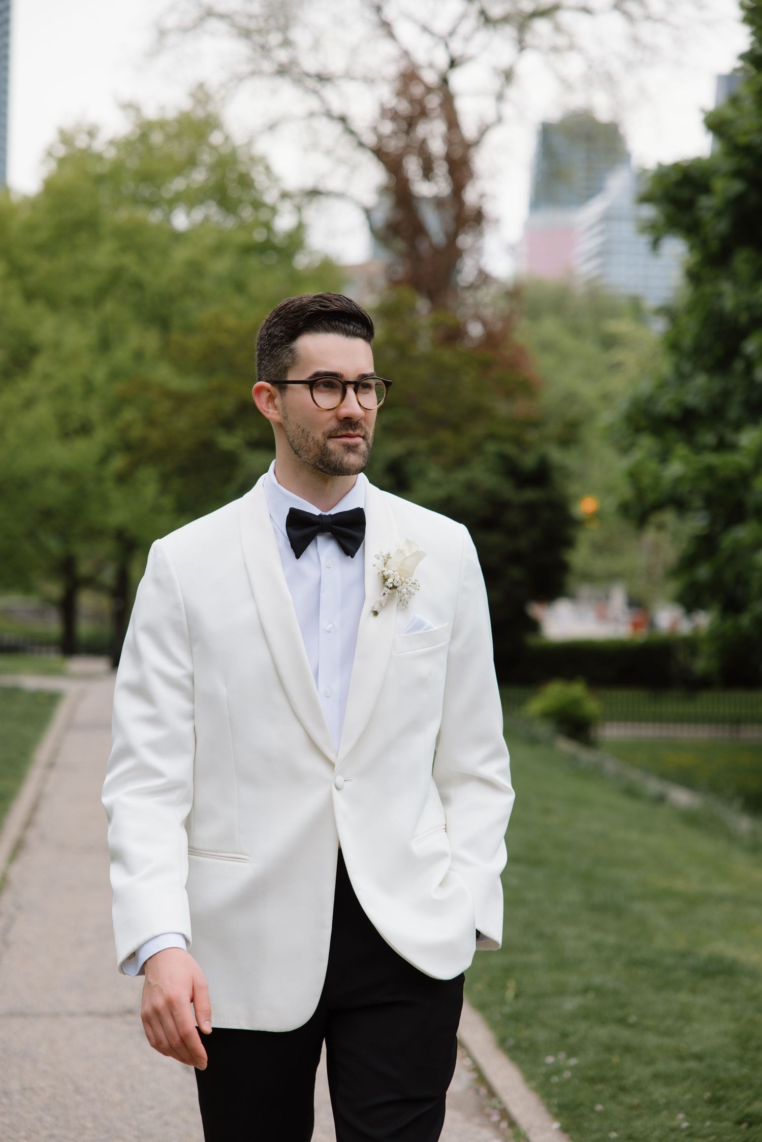 A groom in a classic white dinner jacket and black bow tie poses outdoors in an urban park setting during a wedding.