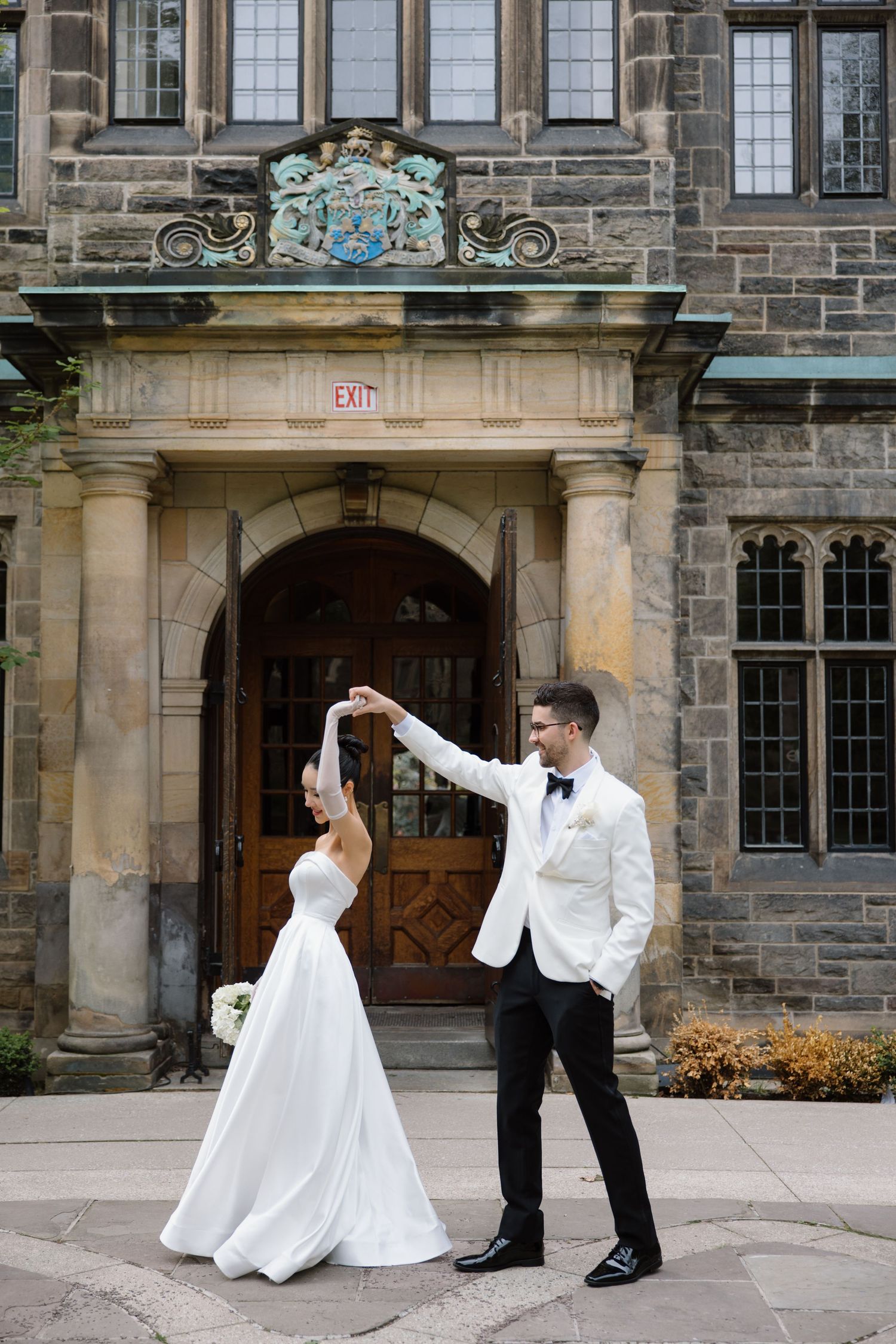 Couple dances joyfully in formal attire outside historic stone building entrance with ornate architectural details.