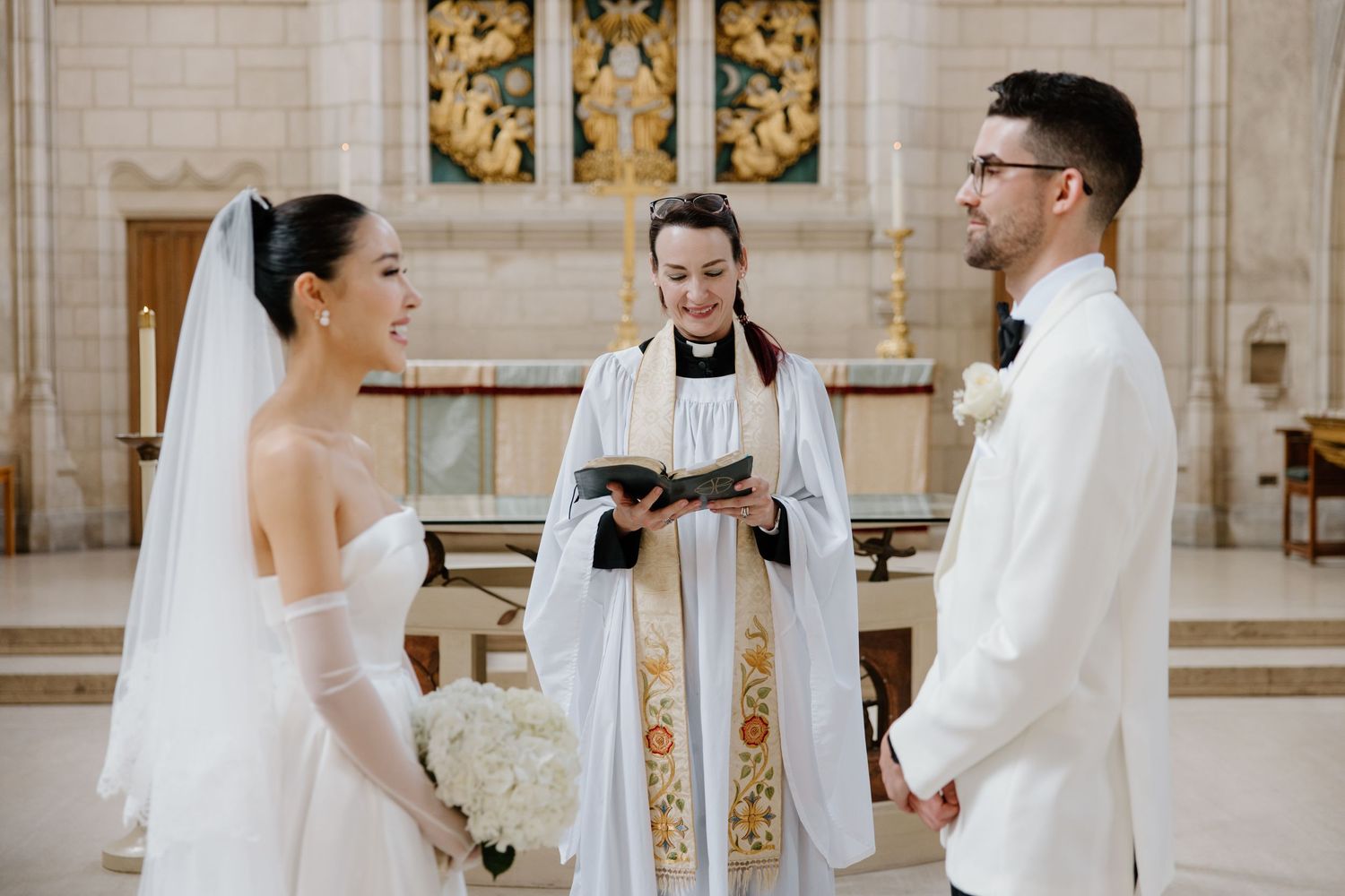 A couple exchanges vows during a wedding ceremony in an elegant church interior decorated with gold details.