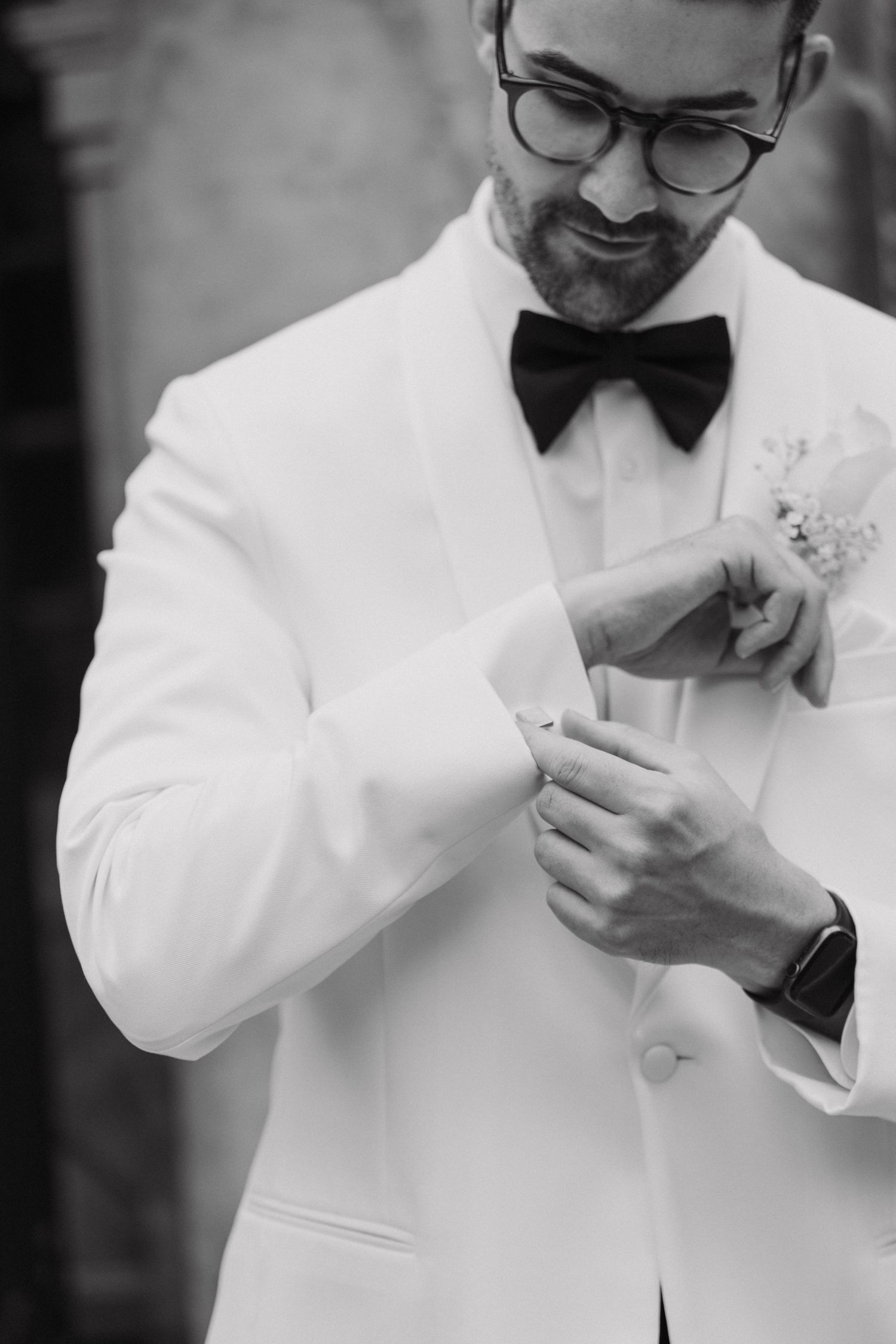 A person in a crisp white tuxedo jacket and black bow tie adjusts their cuff while wearing glasses.