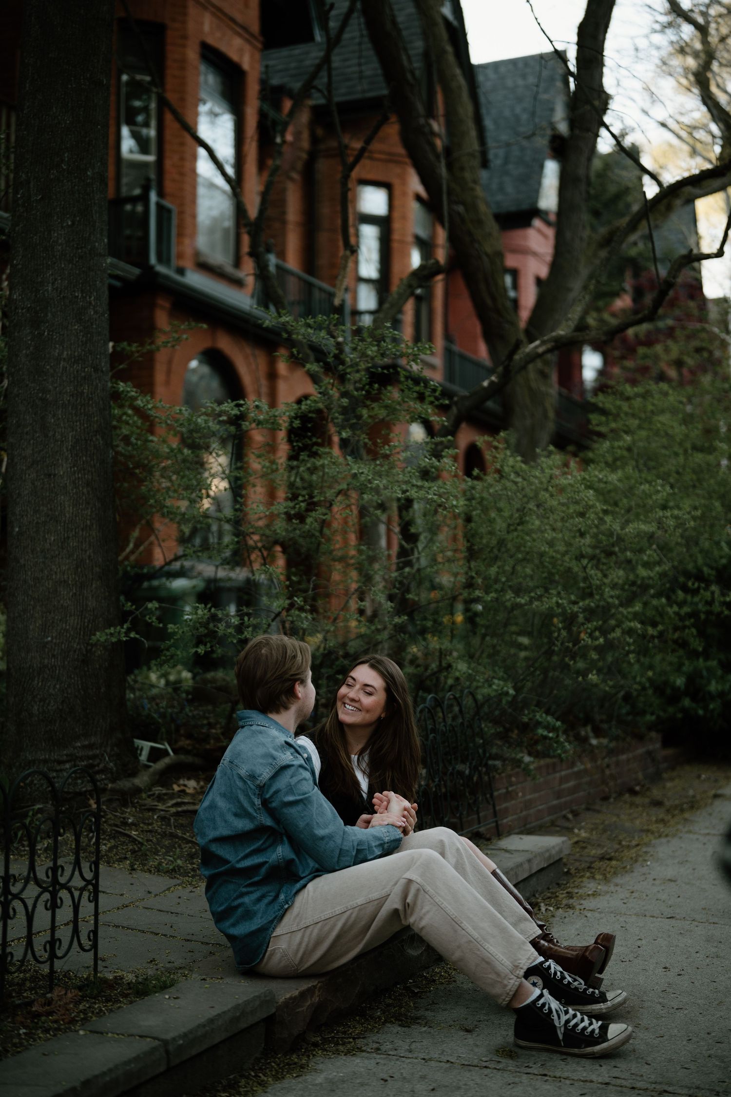 Two people share a tender moment while sitting on a sidewalk near brick townhouses at dusk.