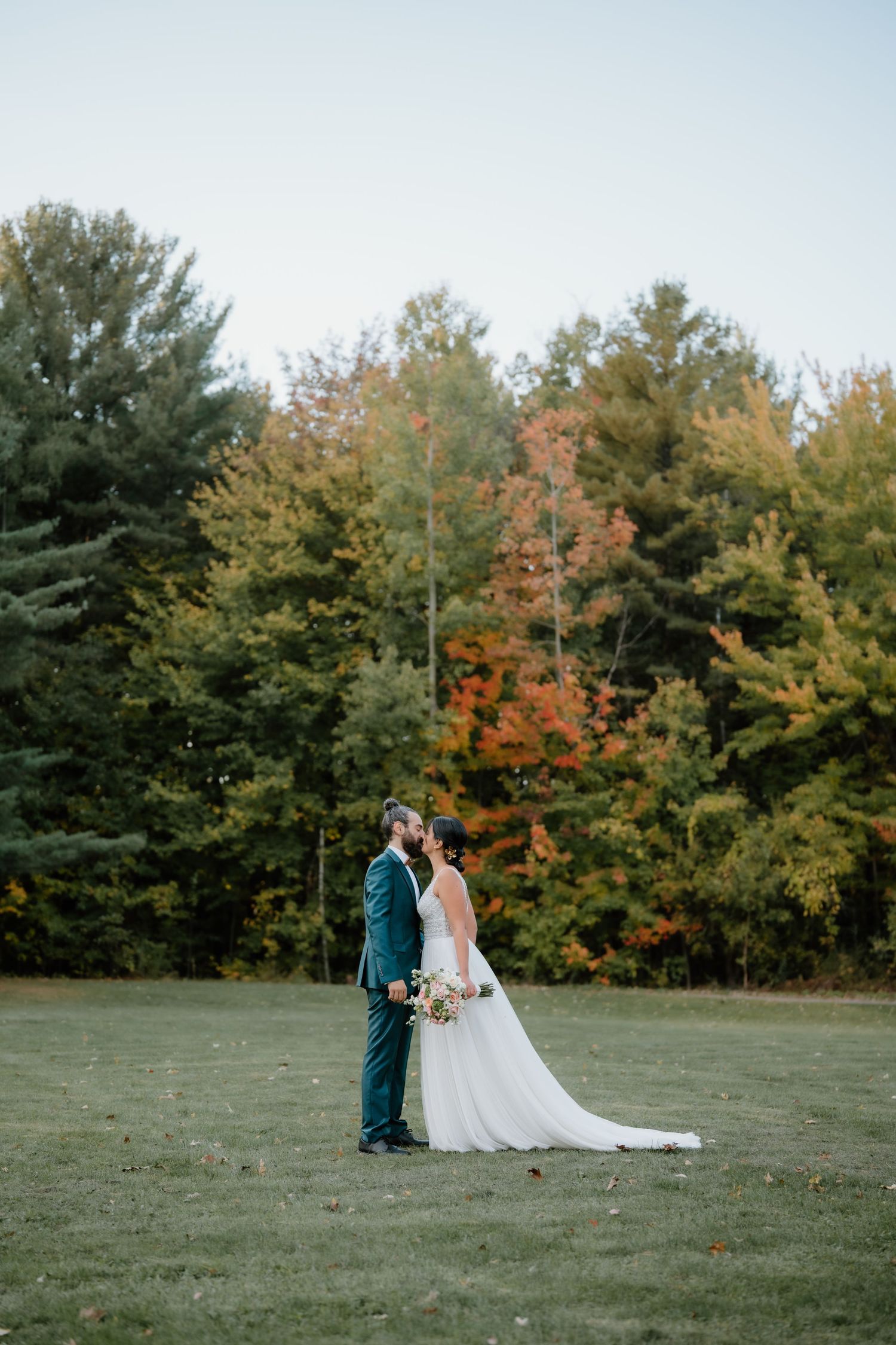 Wedding couple embracing on lawn with autumn trees creating colorful backdrop.