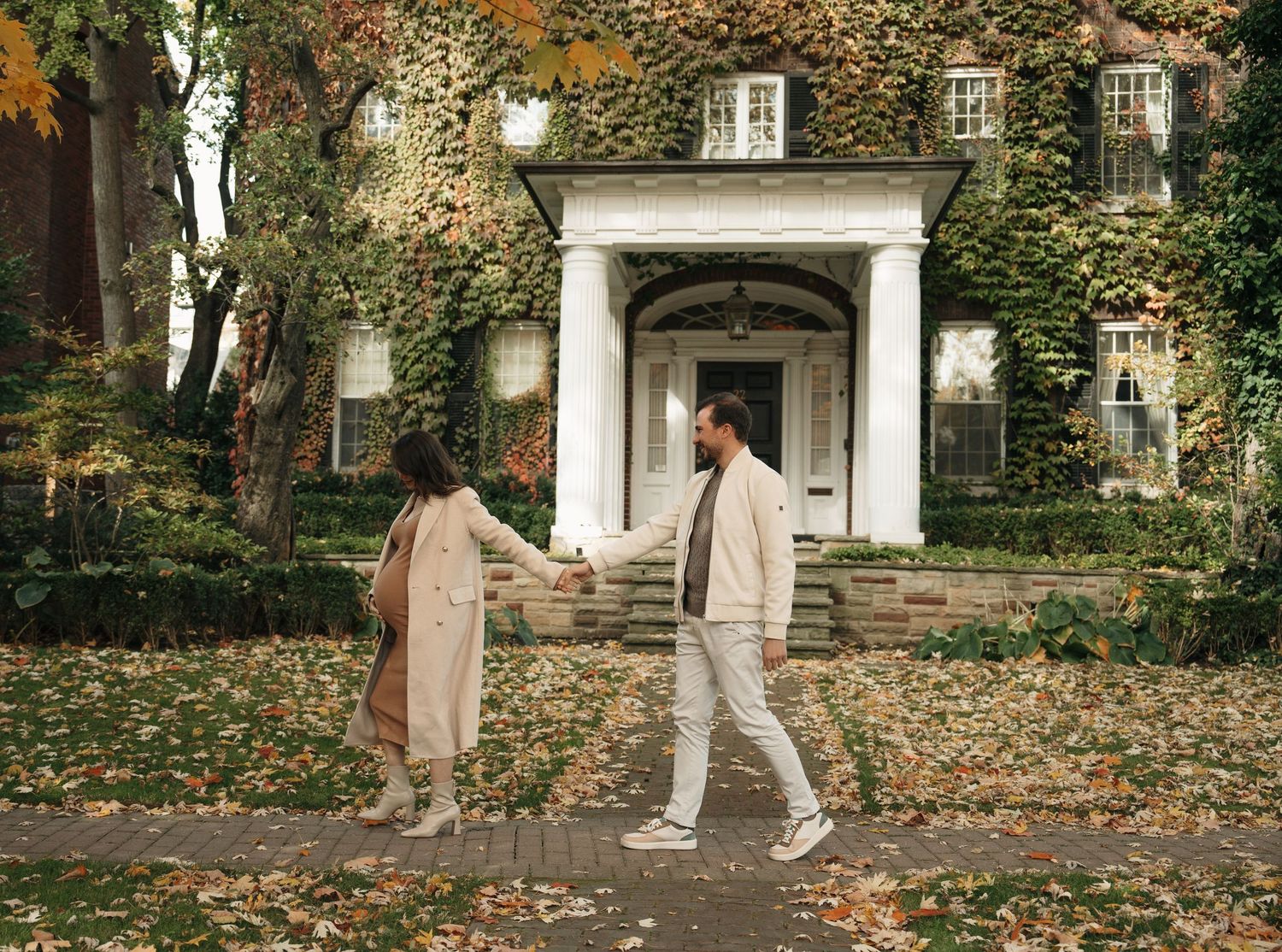 A couple in cream-colored outfits walk through fallen autumn leaves toward an ivy-covered mansion's elegant white entrance.