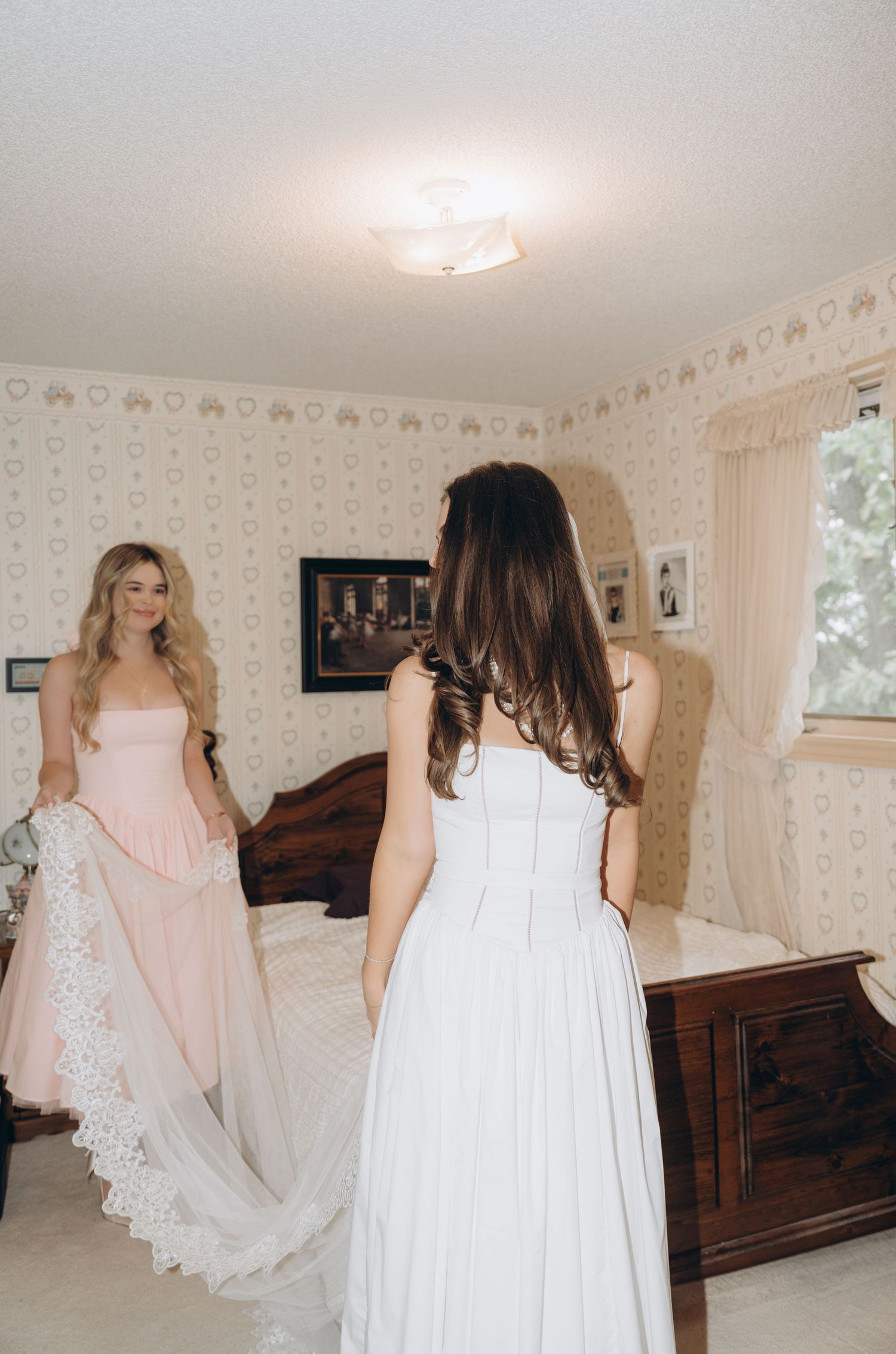 Two women in elegant white dresses stand in a vintage bedroom with floral wallpaper and antique furniture.