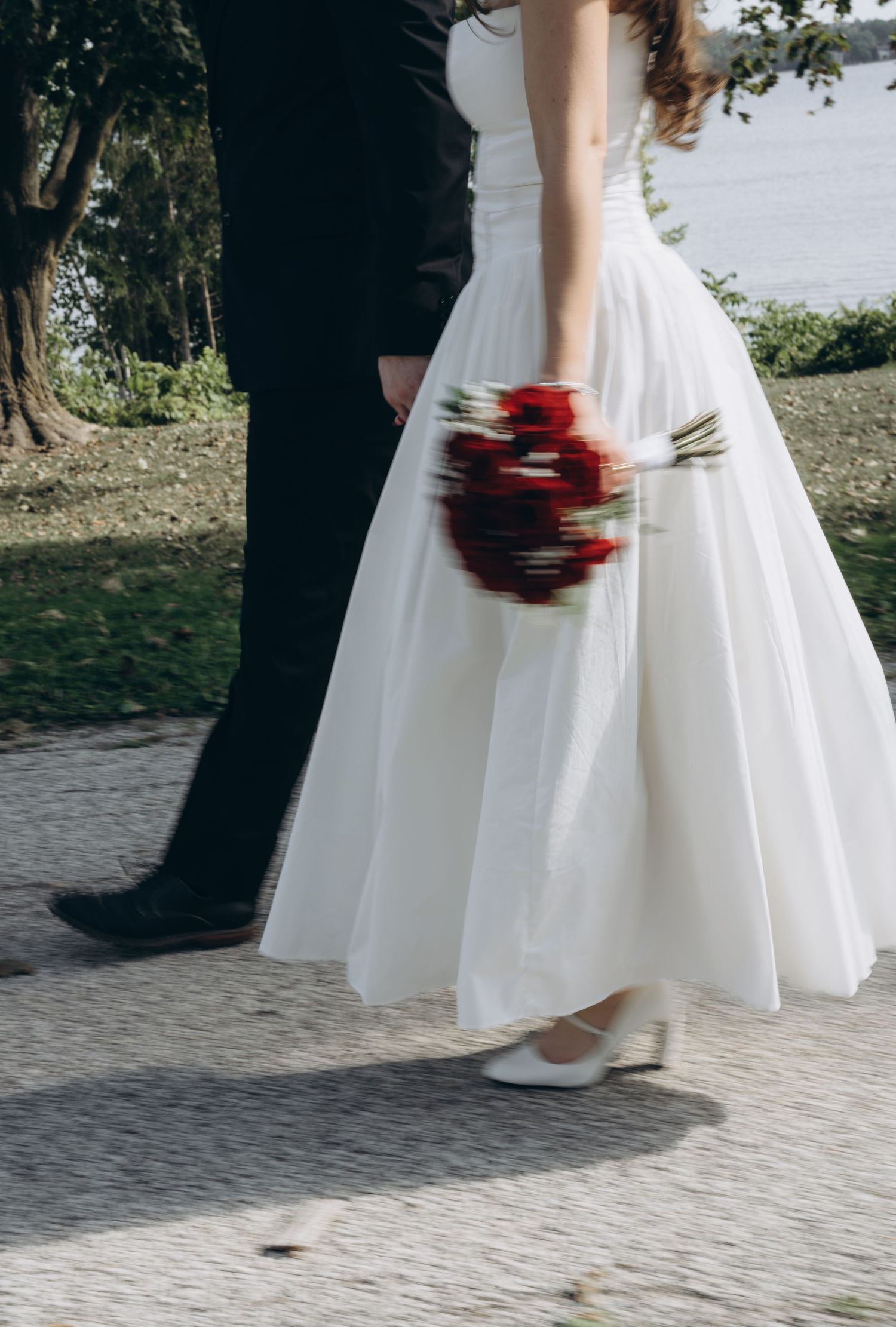 A person in a wedding dress and another in dark attire walking together on a stone path carrying red flowers.