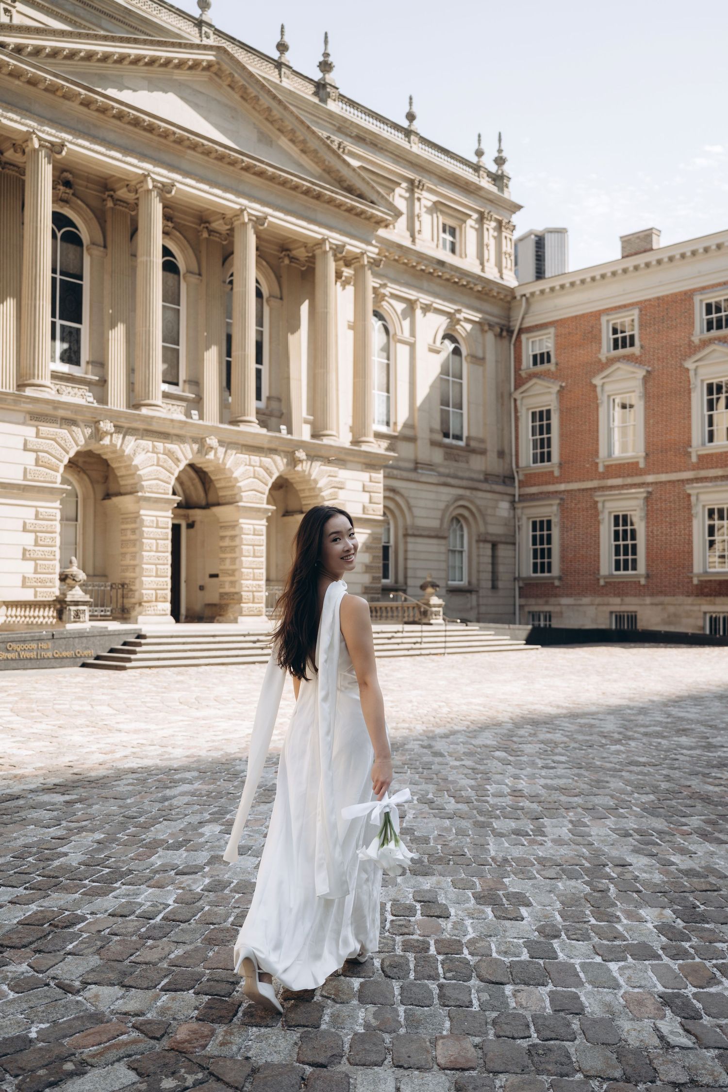 A white-clothed figure walks across a grand cobblestone courtyard surrounded by neoclassical architecture.
