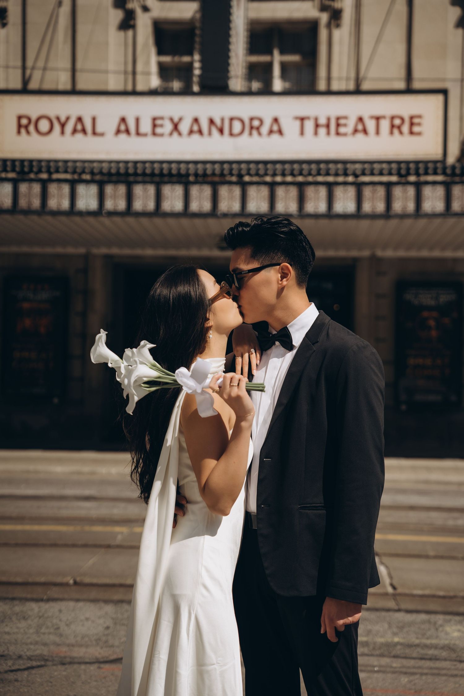A couple shares a romantic kiss outside the Royal Alexandra Theatre, with the bride in white and groom in a tuxedo.