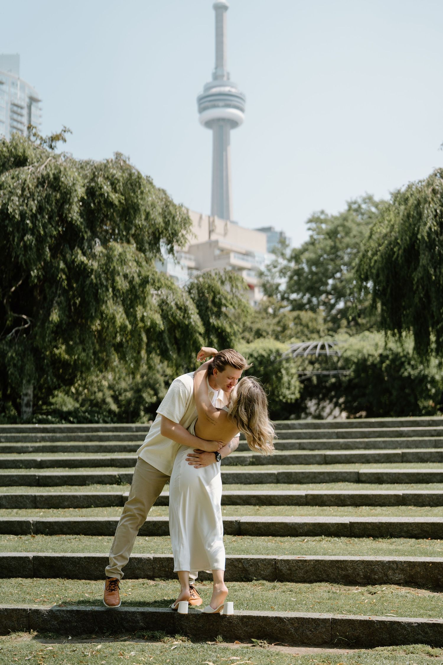 A romantic outdoor moment unfolds on stone steps in a sunlit park with Toronto's CN Tower visible in the background.