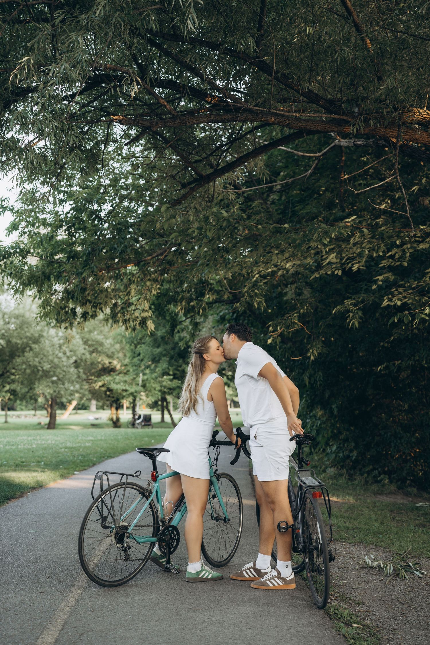 A couple in white summer outfits share a romantic moment with their bicycles on a tree-lined path in a park.