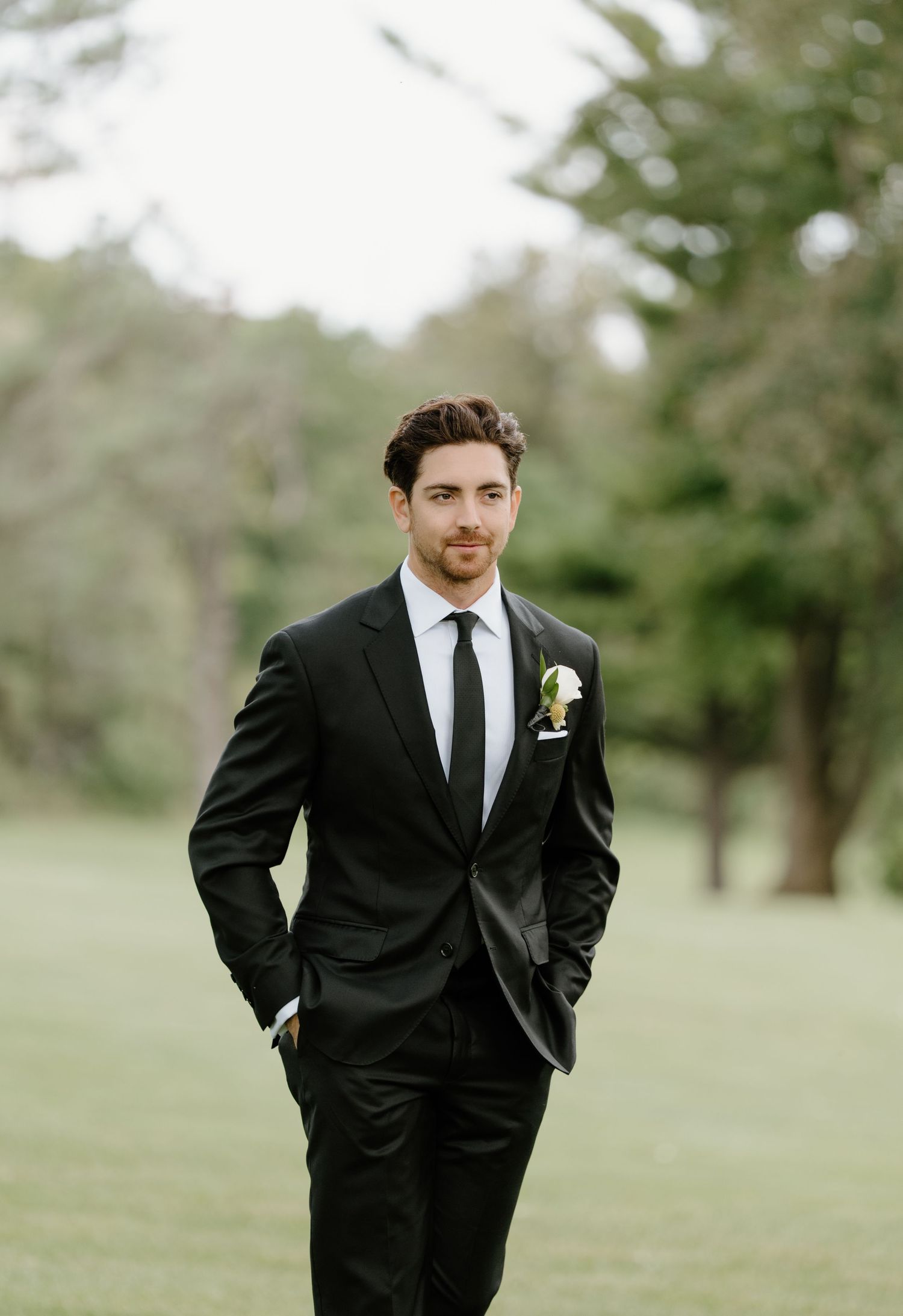 A groom in a classic black tuxedo stands in a grassy outdoor setting with a boutonniere pinned to his lapel.