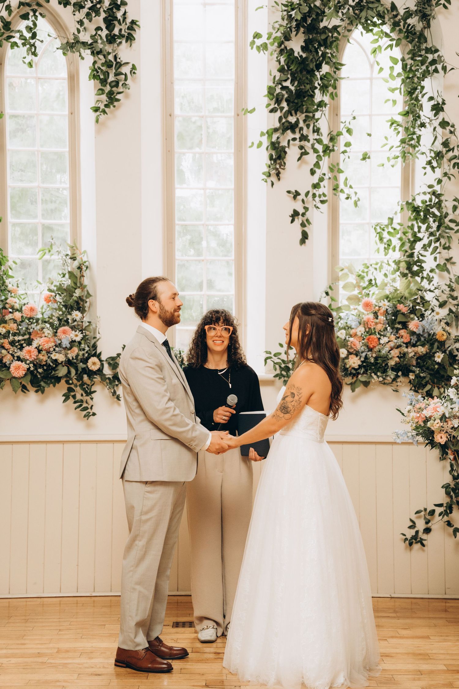 A wedding ceremony takes place in a bright room decorated with cascading greenery and peach flowers by the windows.