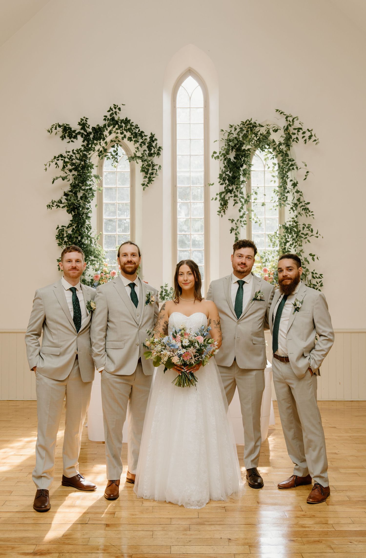 Wedding party poses in light gray suits and white dress inside bright minimalist venue with arched windows and greenery.