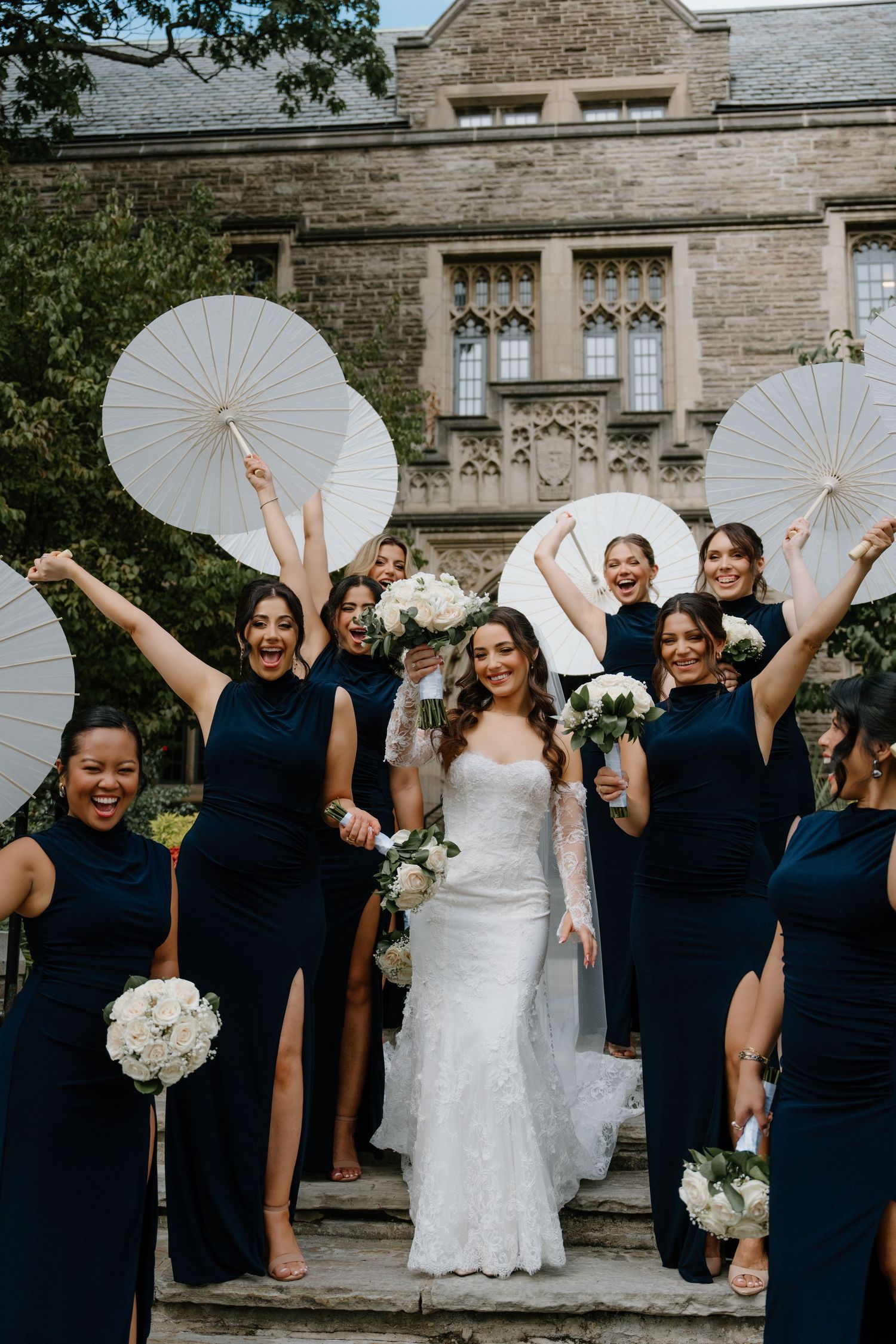 Wedding party in navy blue dresses holding white parasols poses joyfully outside stone church building.