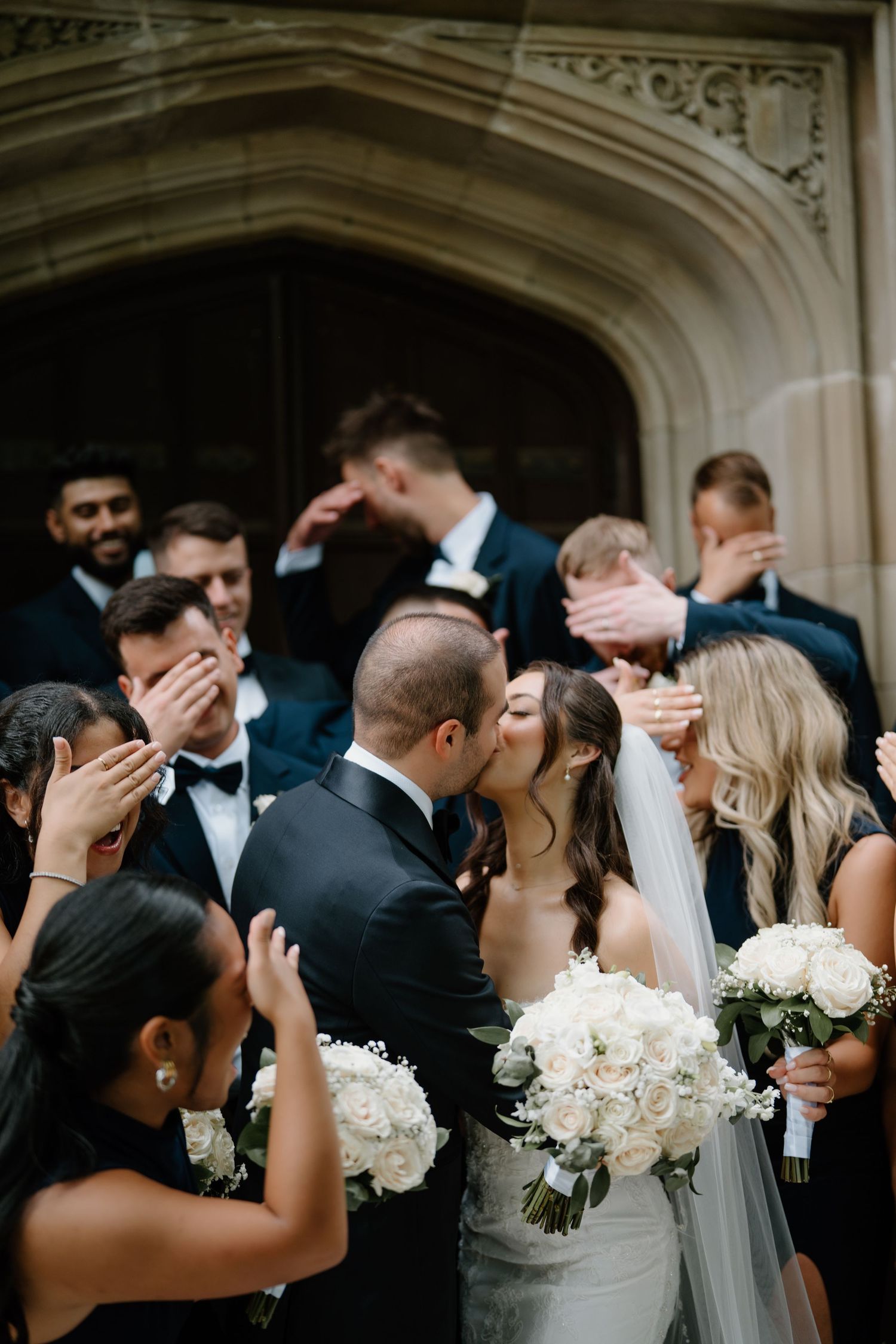 Wedding party members in formal attire share romantic kisses while holding white floral bouquets outside an elegant venue.