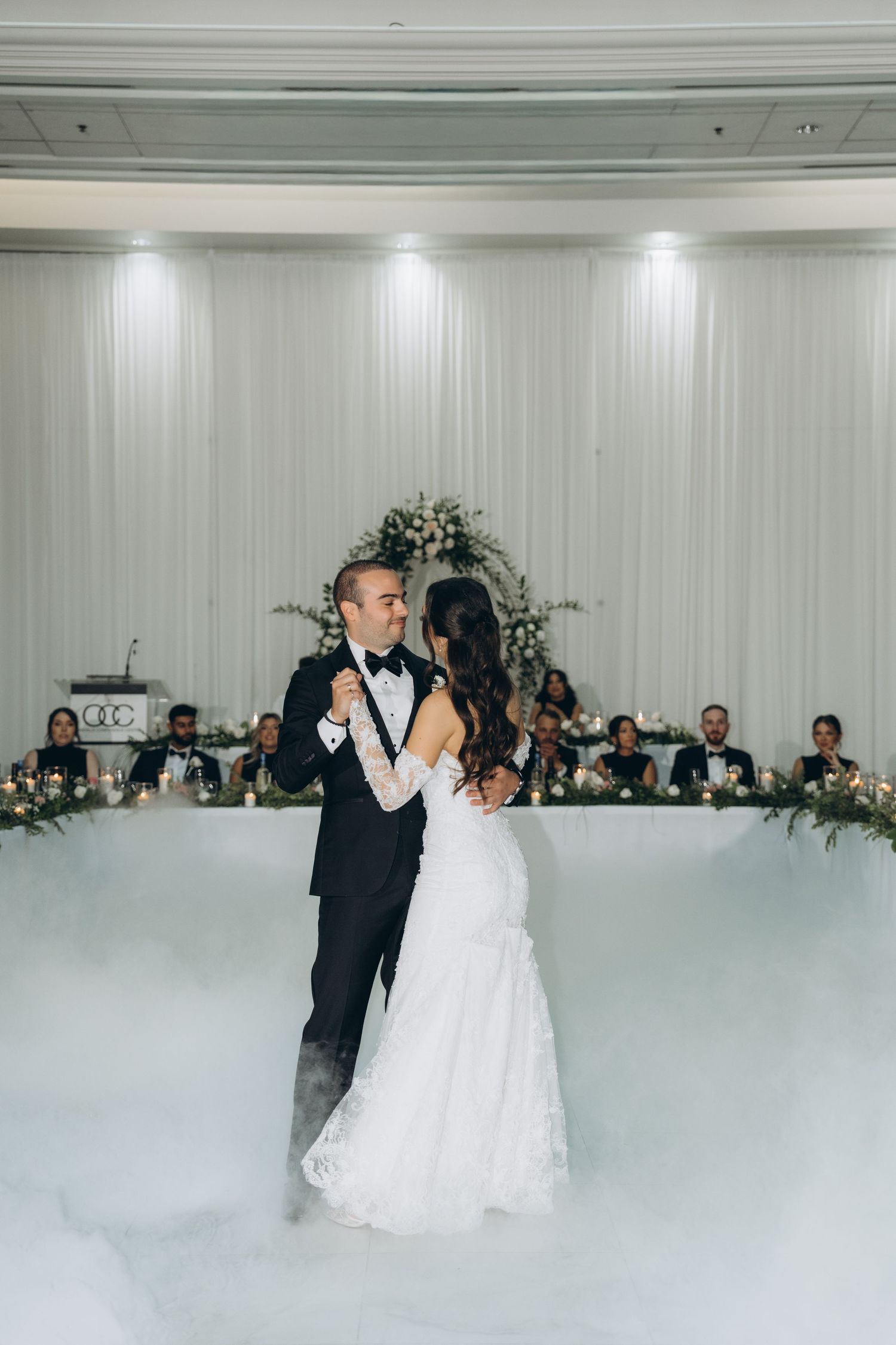 Newlyweds share their first dance on a white dance floor at an elegant wedding reception with guests watching.