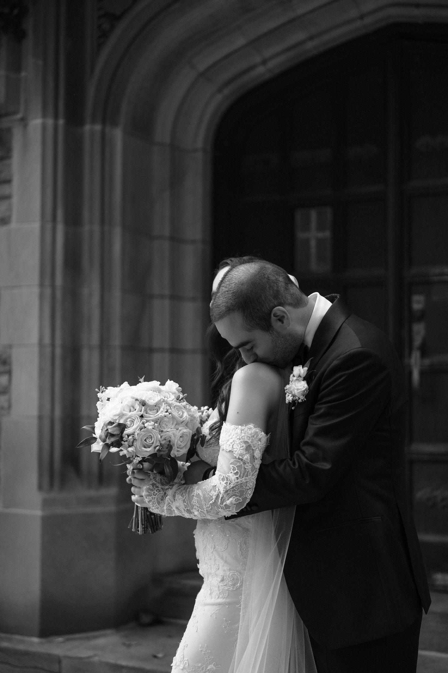 A black and white wedding photograph showing an intimate moment between newlyweds against an architectural backdrop.