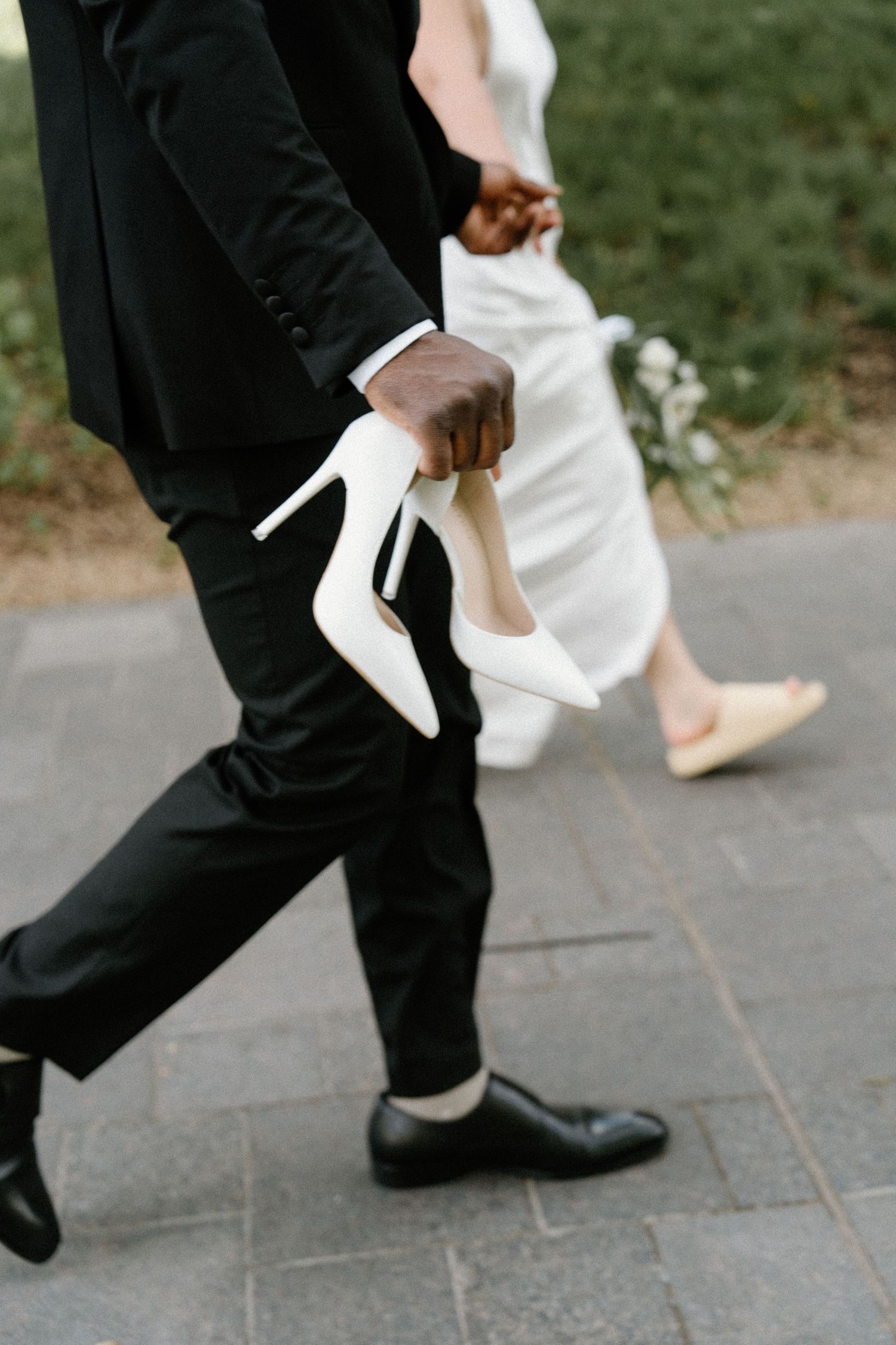 A person in a black suit helps someone remove white high heeled shoes while standing on pavement.