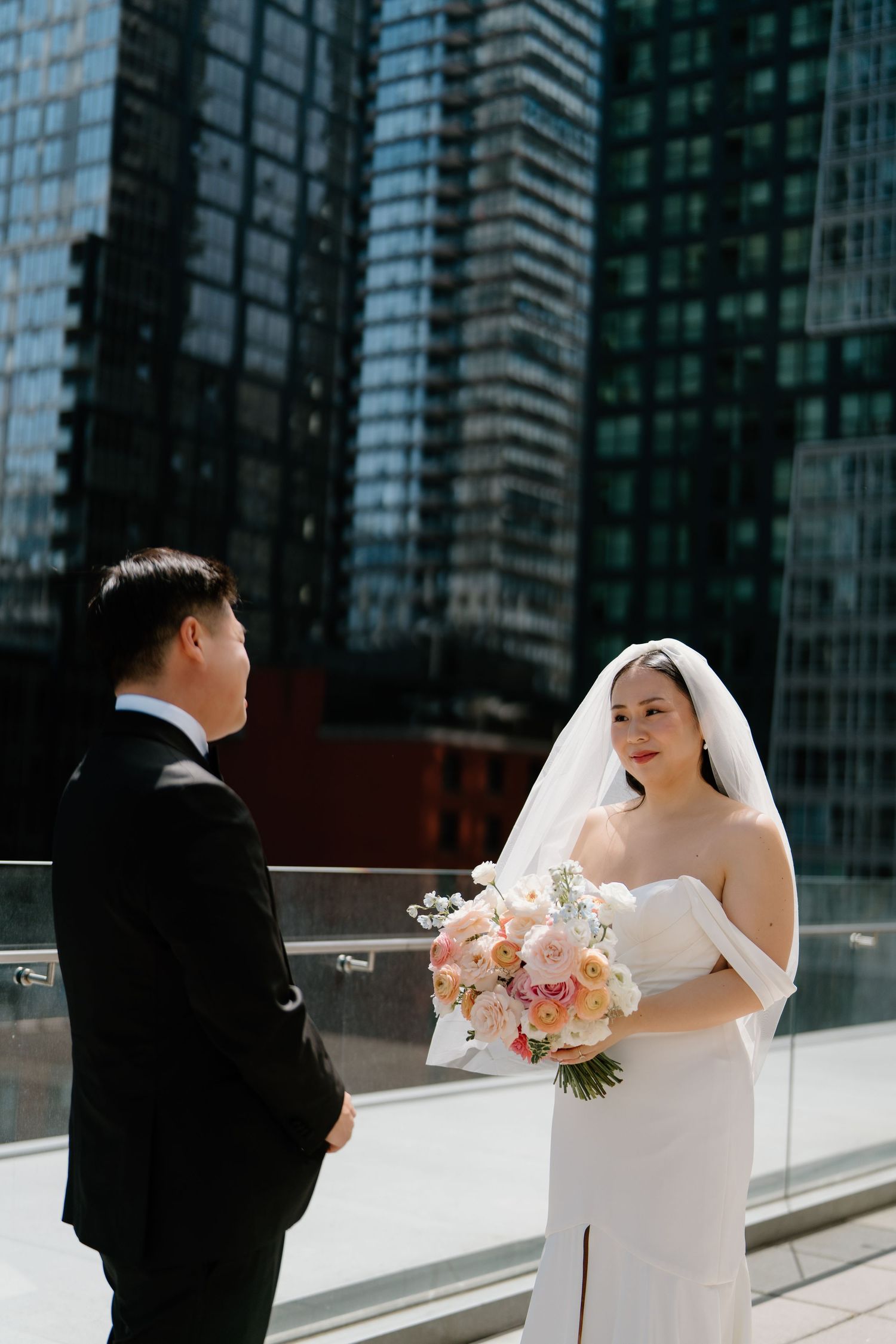 A couple in wedding attire posing outdoors with modern glass skyscrapers in the background.