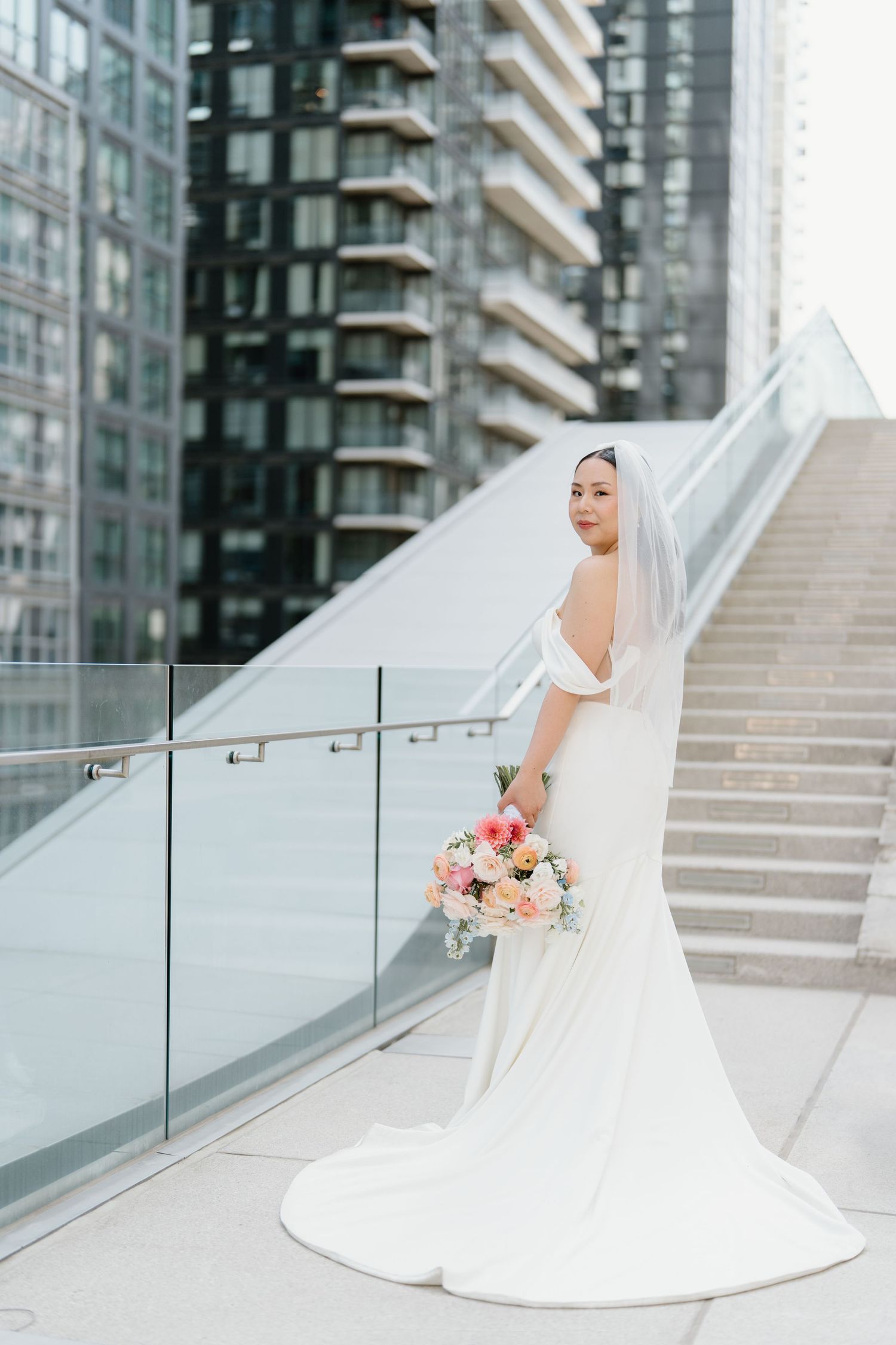 Elegant bride in white gown holds colorful bouquet while standing on urban rooftop terrace with glass railings and stairs.