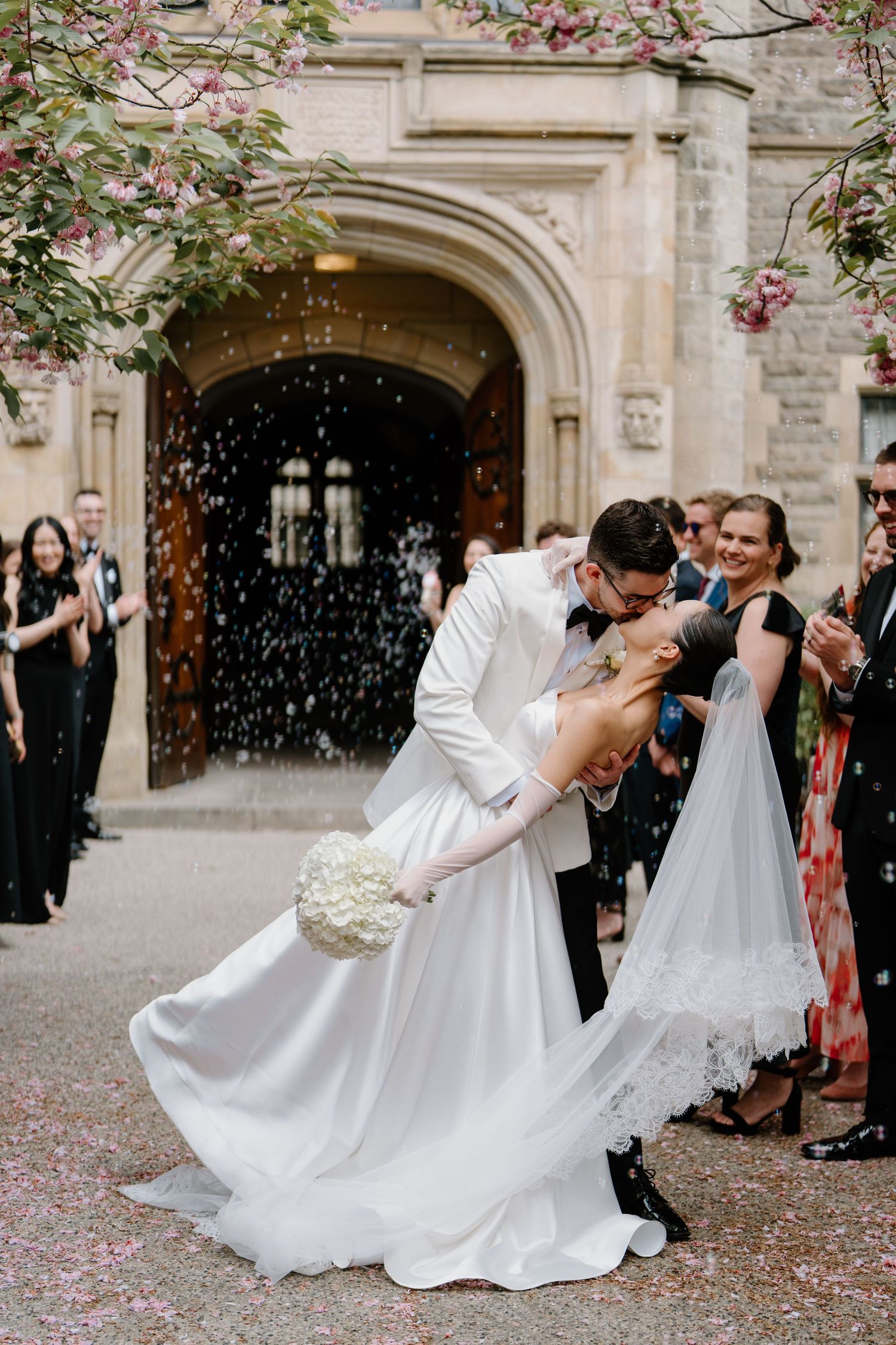 A romantic wedding scene with confetti falling while the couple shares a traditional dip kiss outside a grand stone archway.