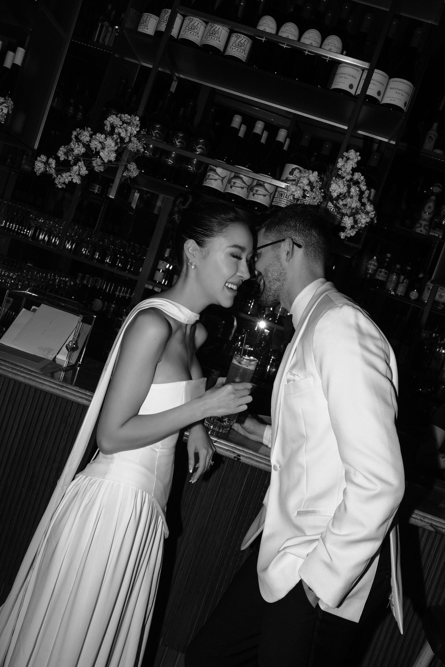 Black and white photo of romantic couple laughing together at bar, dressed in formal white wedding attire.