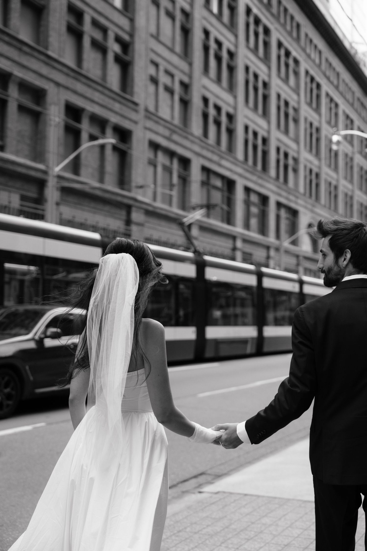 A black and white photo of a couple in wedding attire walking along an urban street with a streetcar visible.