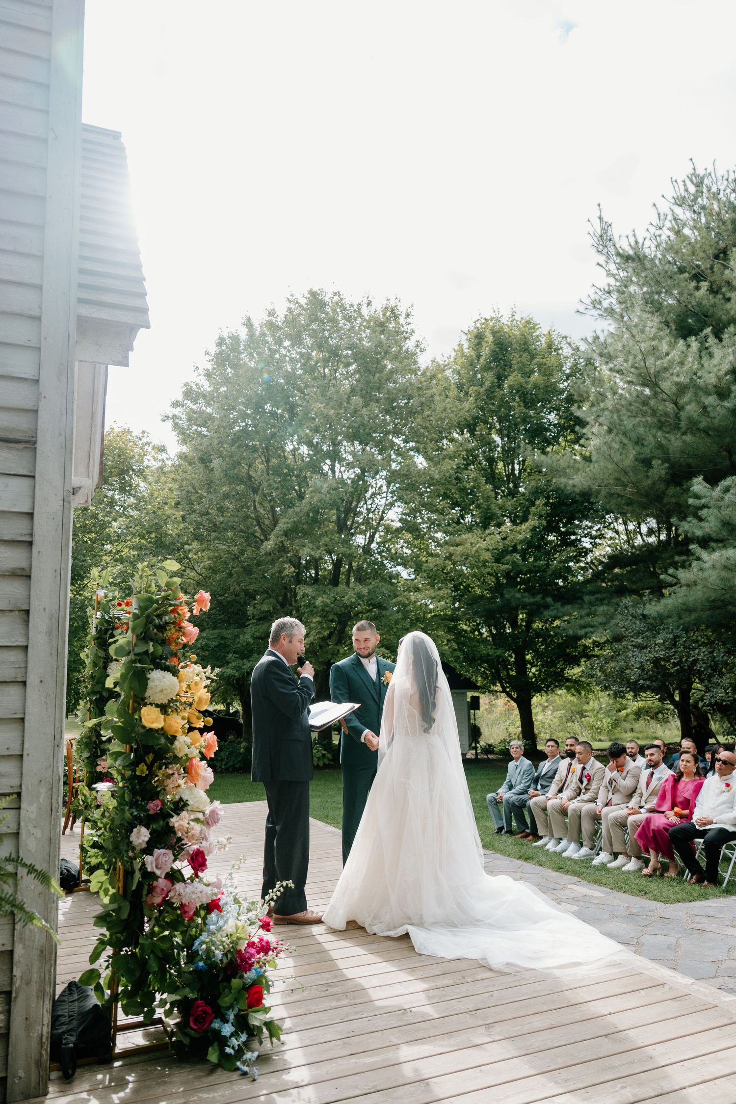 A wedding ceremony takes place on a white wooden deck adorned with floral arrangements and guests seated outdoors.