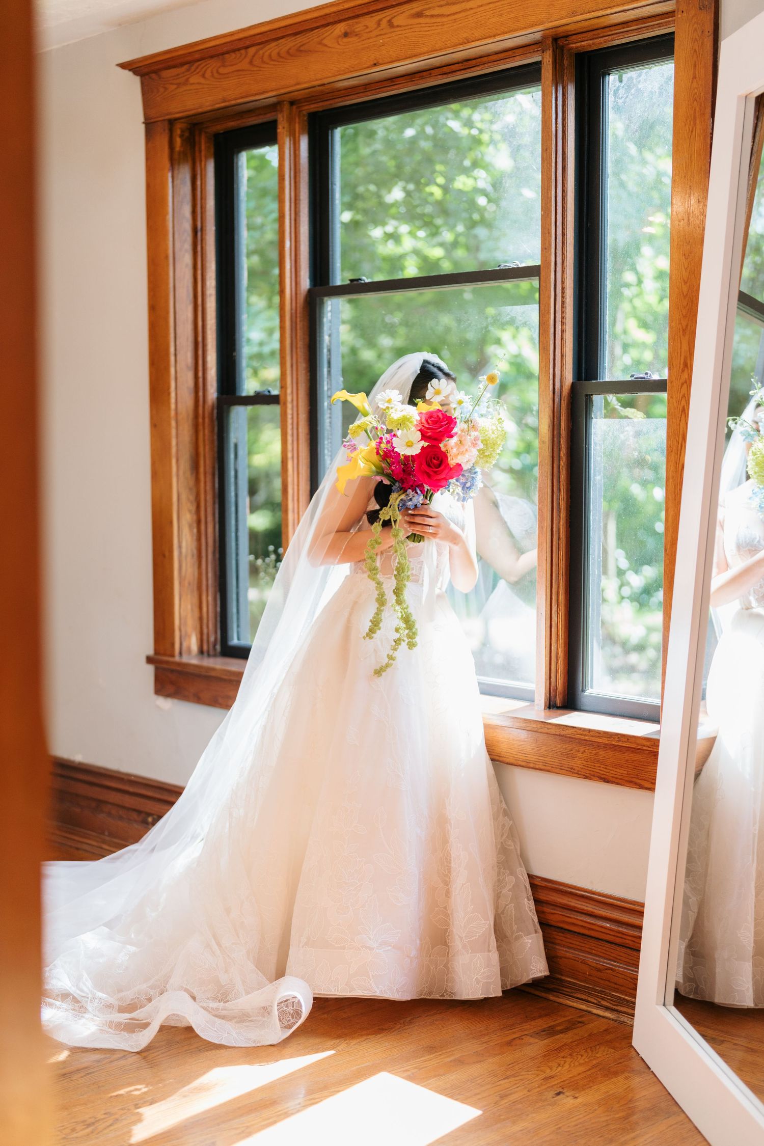A white wedding dress with flowing train and floral bouquet is displayed in front of sunlit windows in a wooden room.