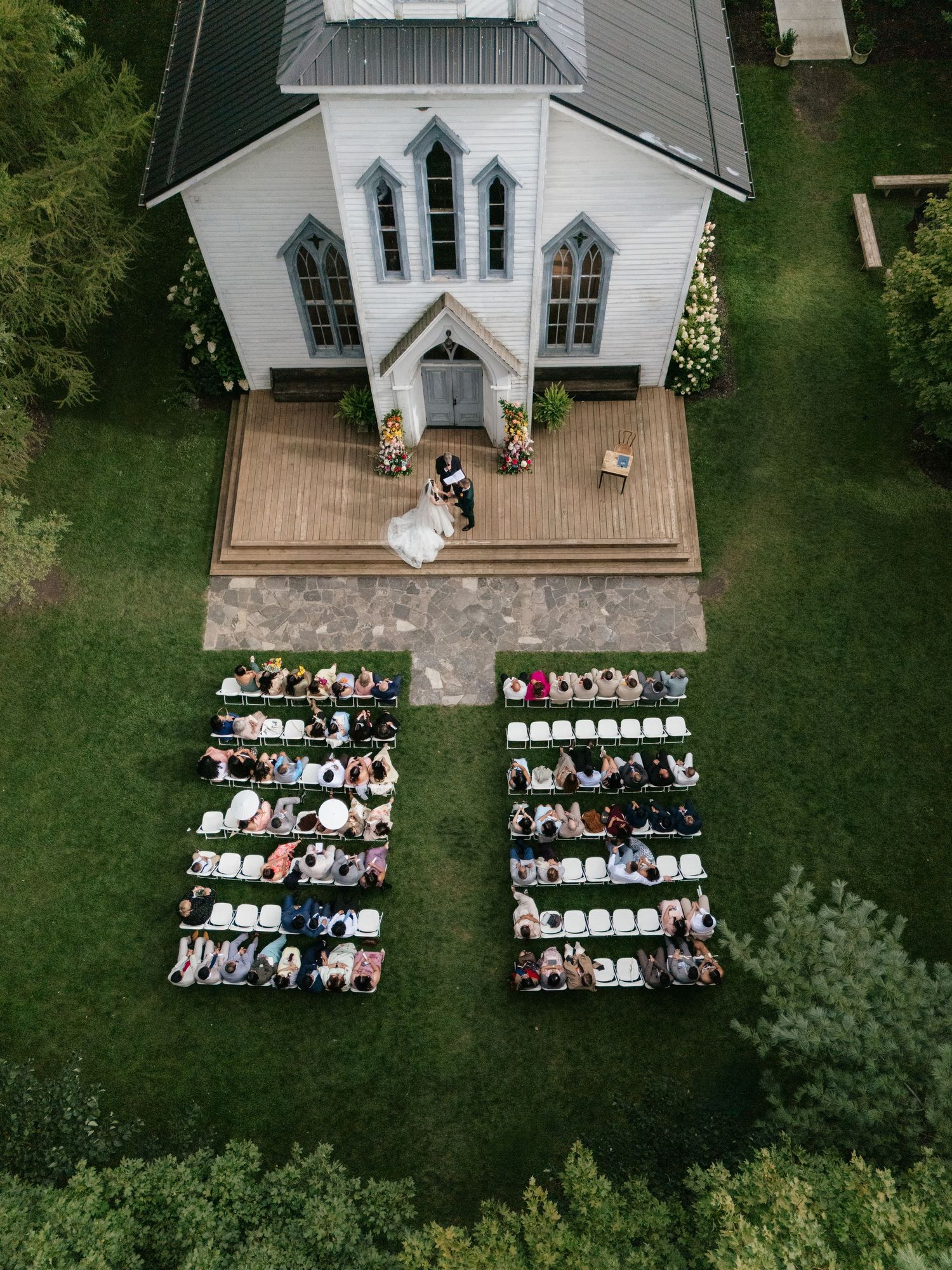 Bird's eye view of wedding guests seated in white chairs outside a charming white chapel.