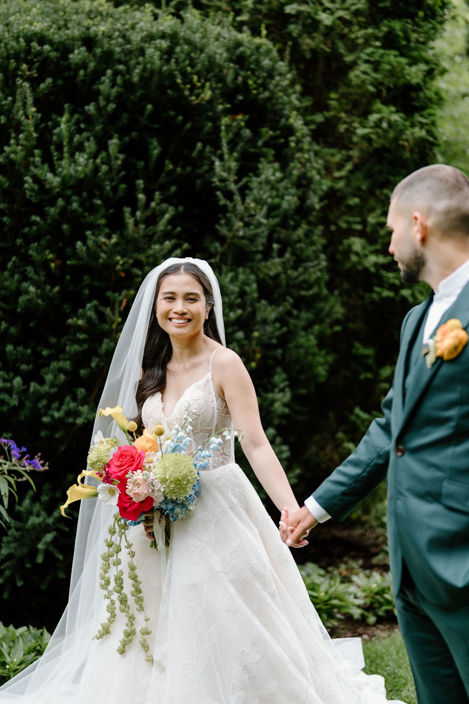 Bride in flowing white gown holds vibrant bouquet while walking through garden path.