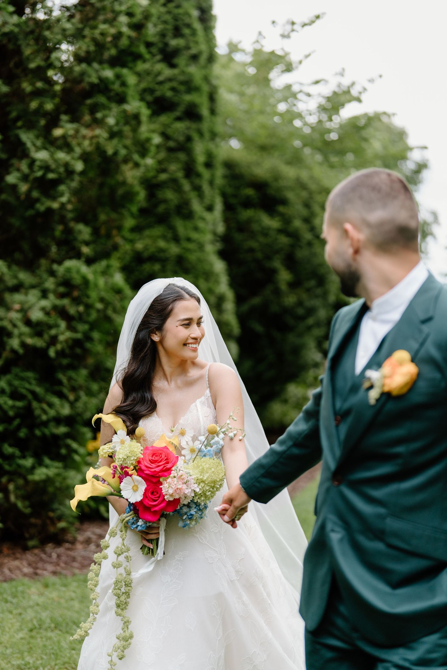 A bride in a white dress holds a vibrant bouquet while walking with someone wearing a dark green suit outdoors.