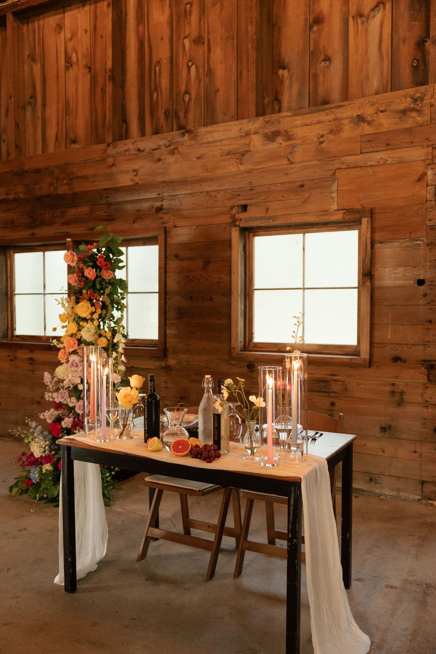 Rustic barn wedding table with white draping, candles, floral arrangements and hanging lanterns against wooden plank walls.