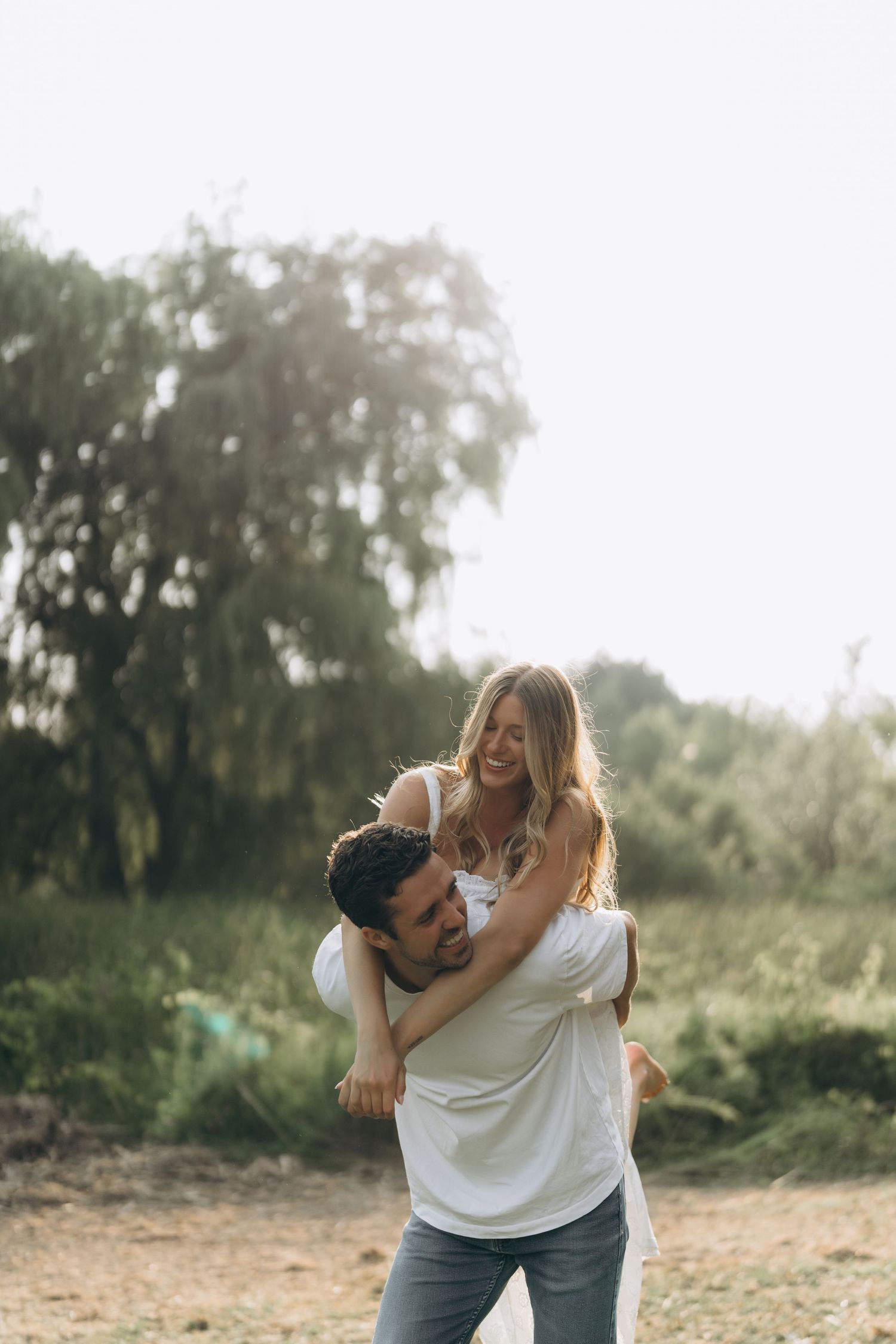 A couple in white shirts and jeans playfully embrace outdoors in natural sunlight during a candid photoshoot.