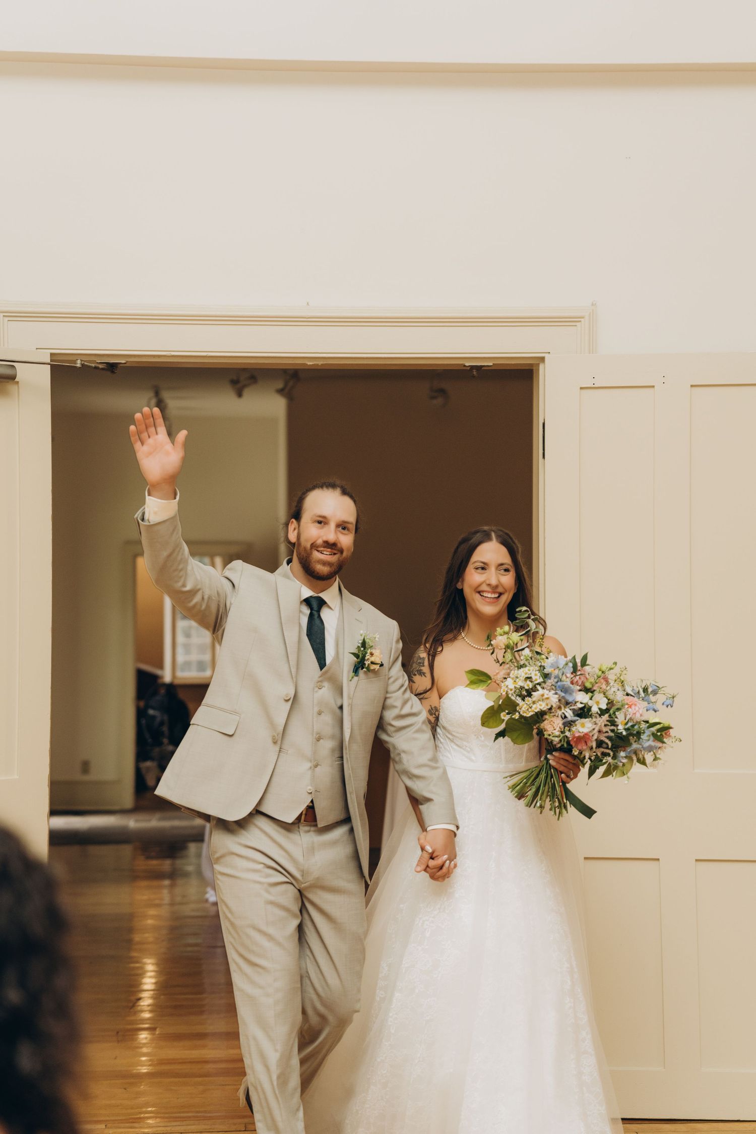 Newlywed couple in light gray suit and white wedding dress wave while entering reception venue.